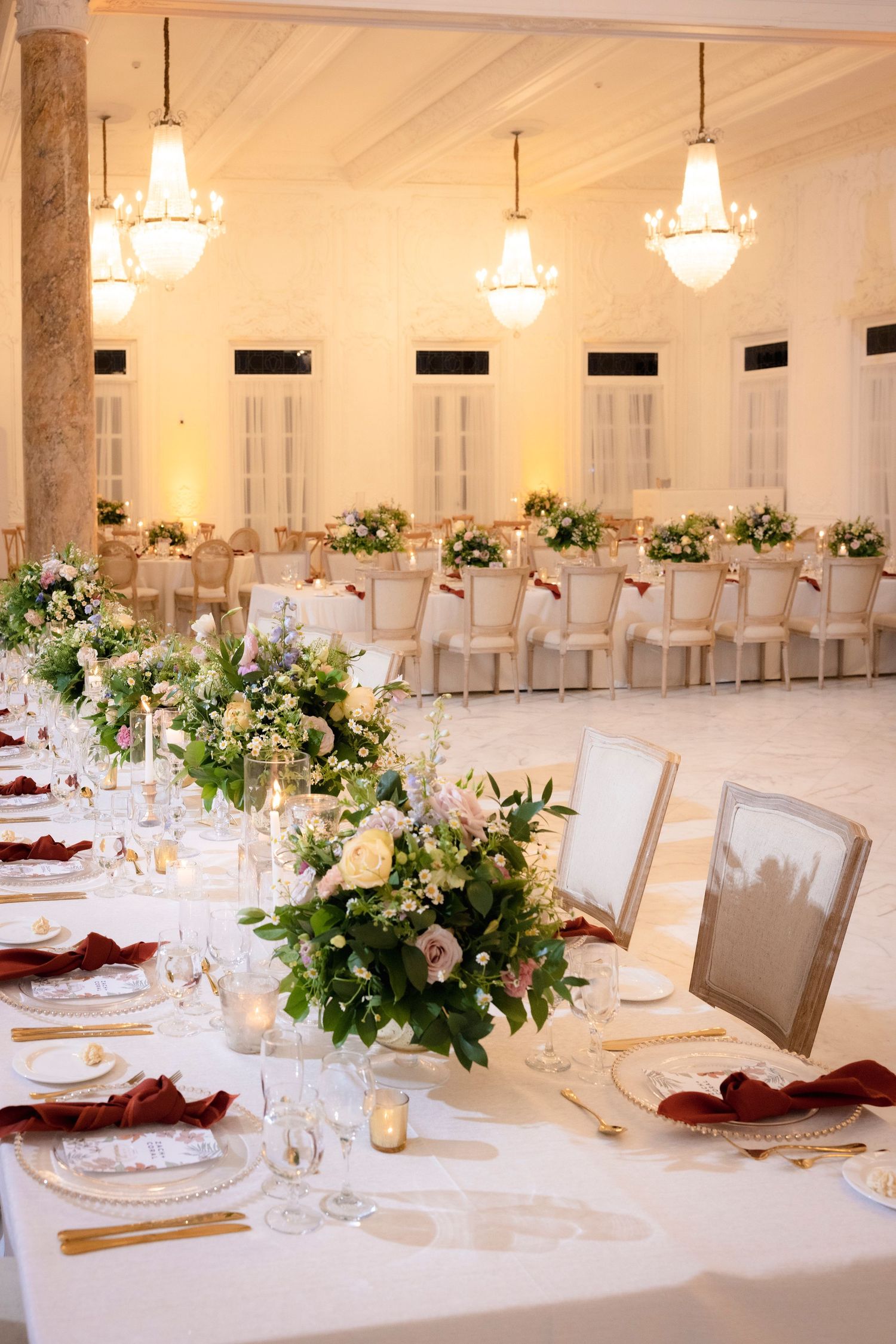 Elegant ballroom wedding reception with white chairs and floral centerpieces under crystal chandeliers.