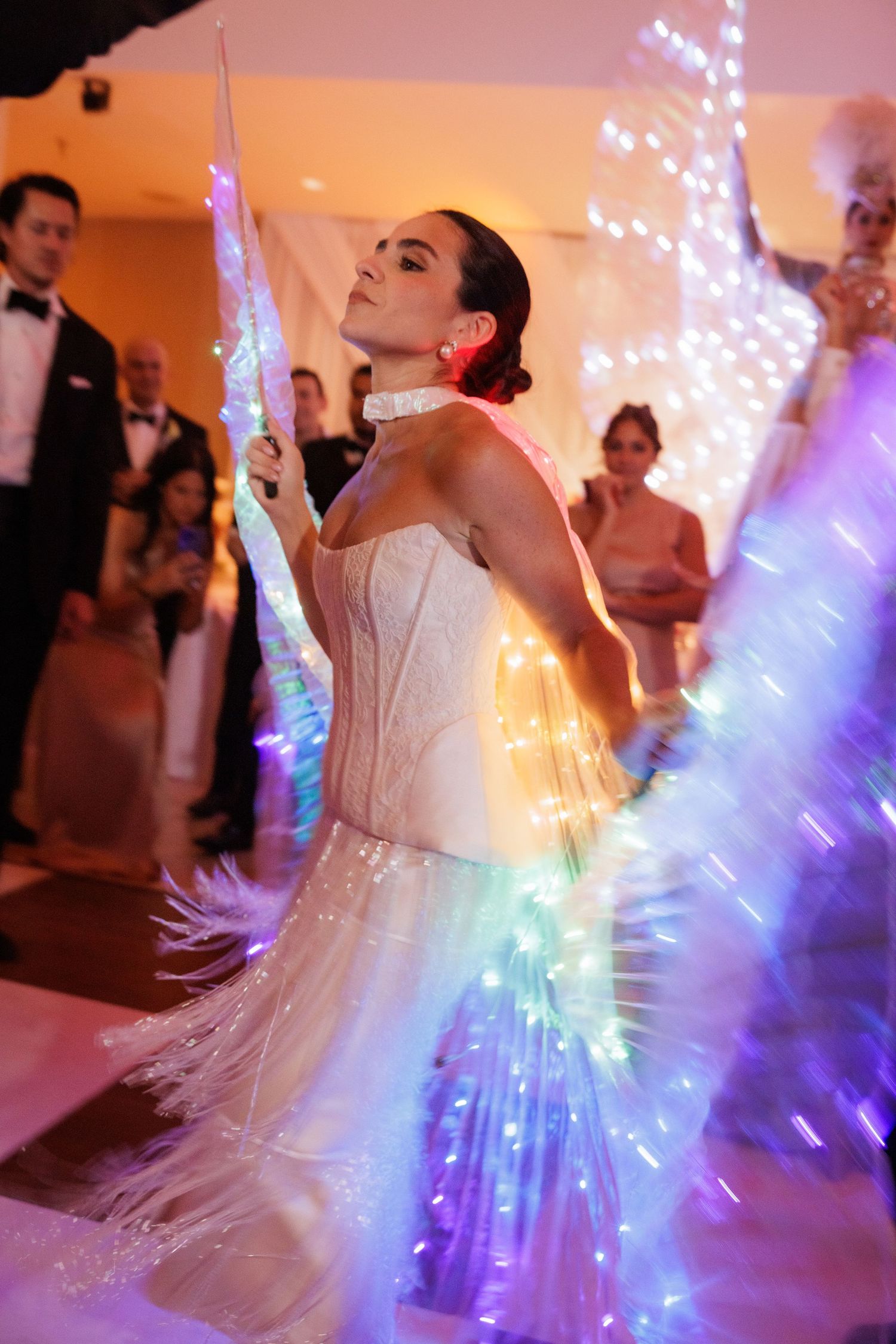 A performer in a white dress creates stunning light patterns with illuminated LED wings at a celebration.