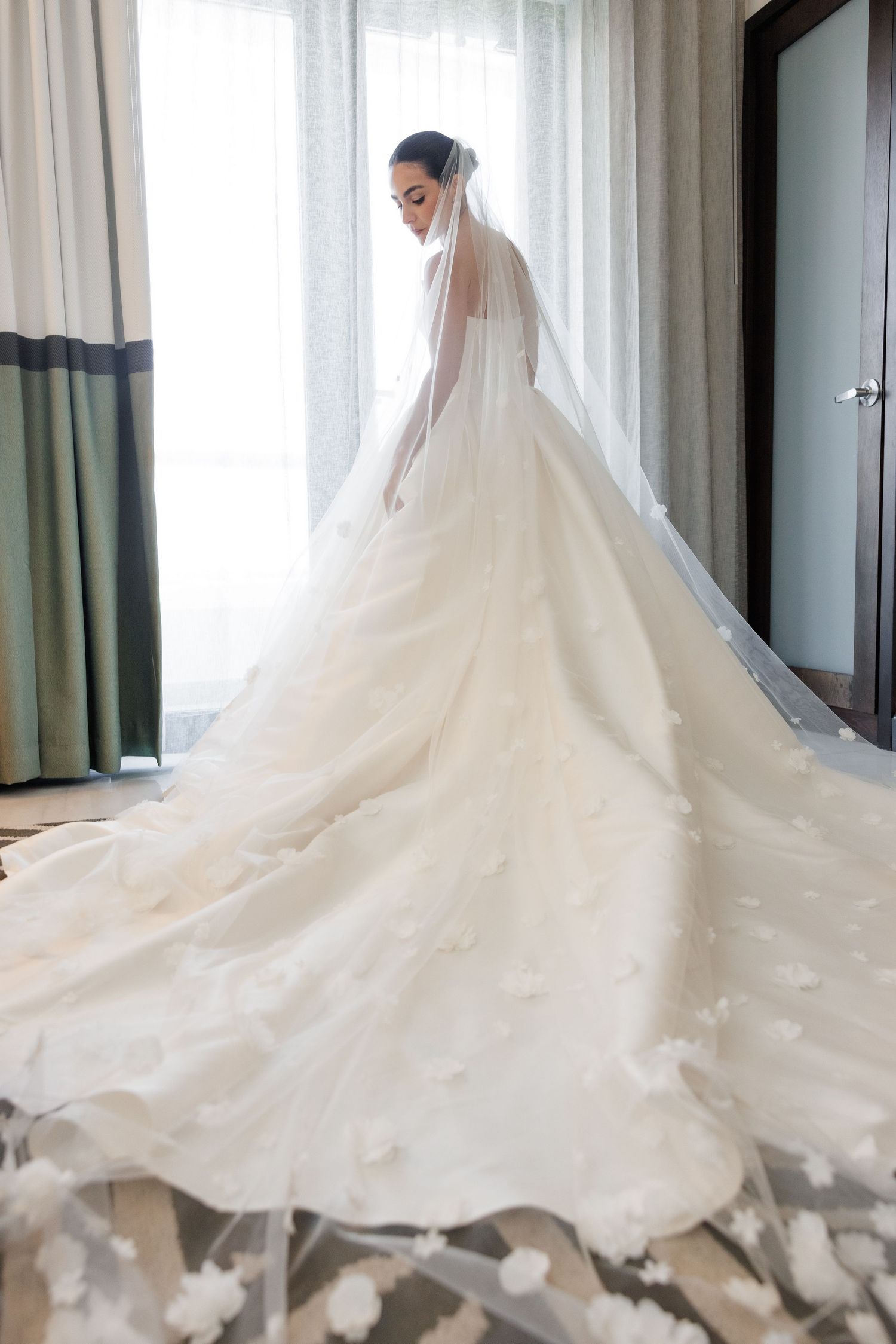 A flowing white wedding dress with a dramatic cathedral-length train spreads elegantly across a light-filled room.