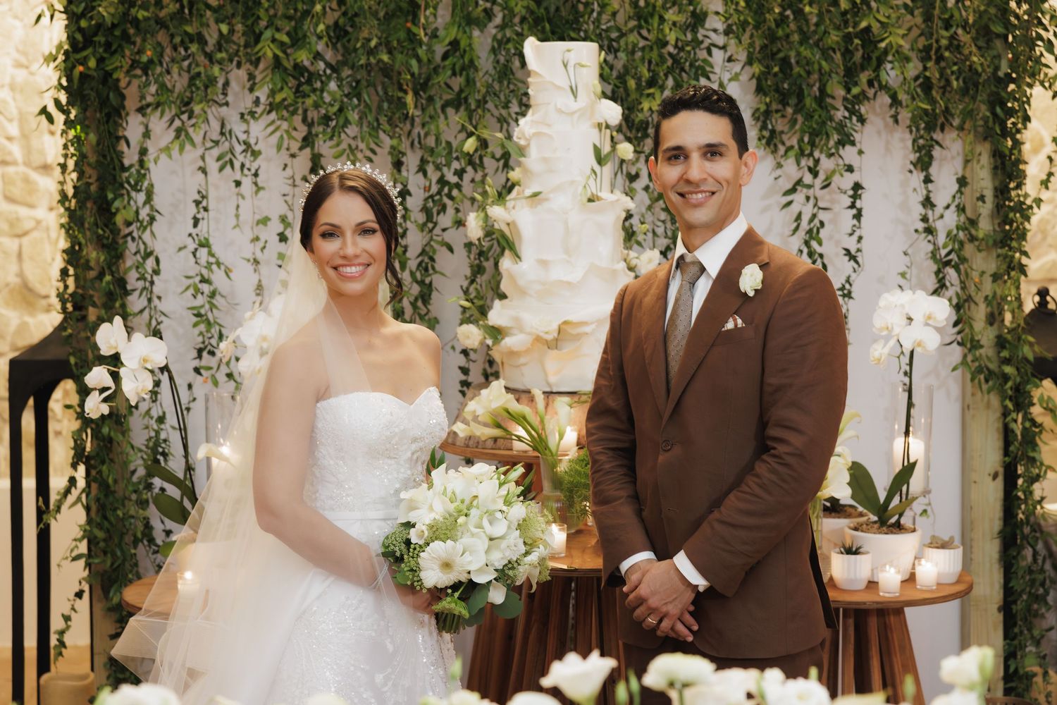 A couple in wedding attire poses together in front of an elegant ivy-covered wall at their wedding ceremony.