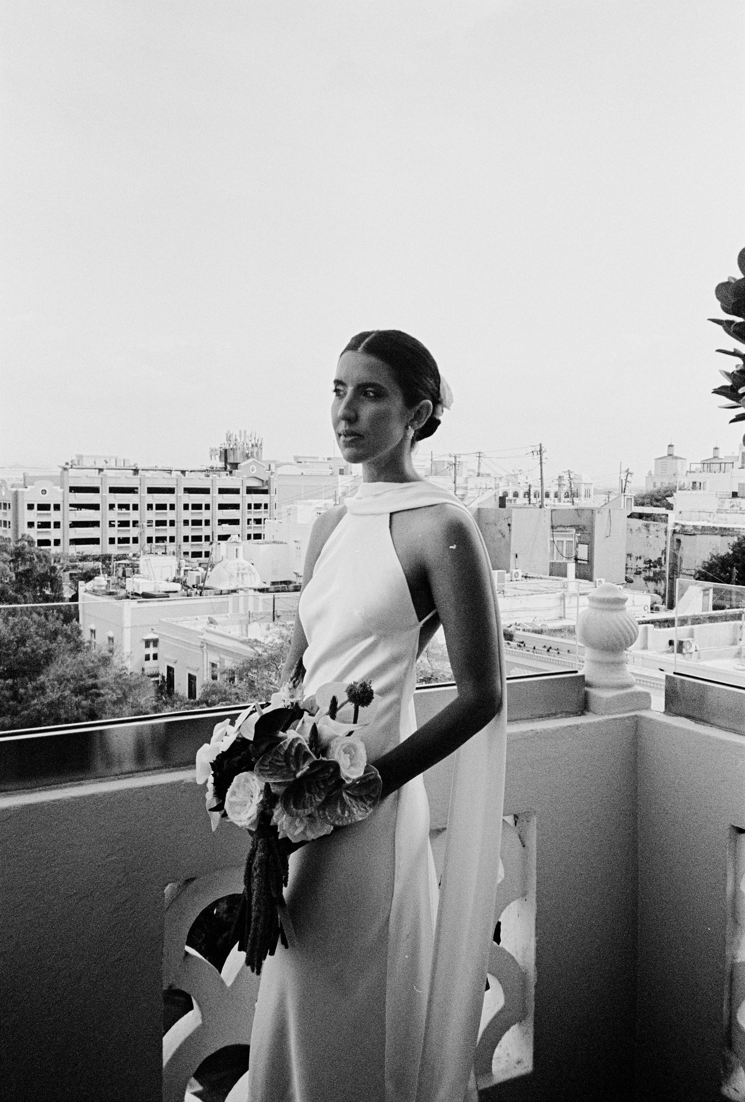 Bride in white halter gown holds bouquet on city rooftop balcony, black and white film photography.