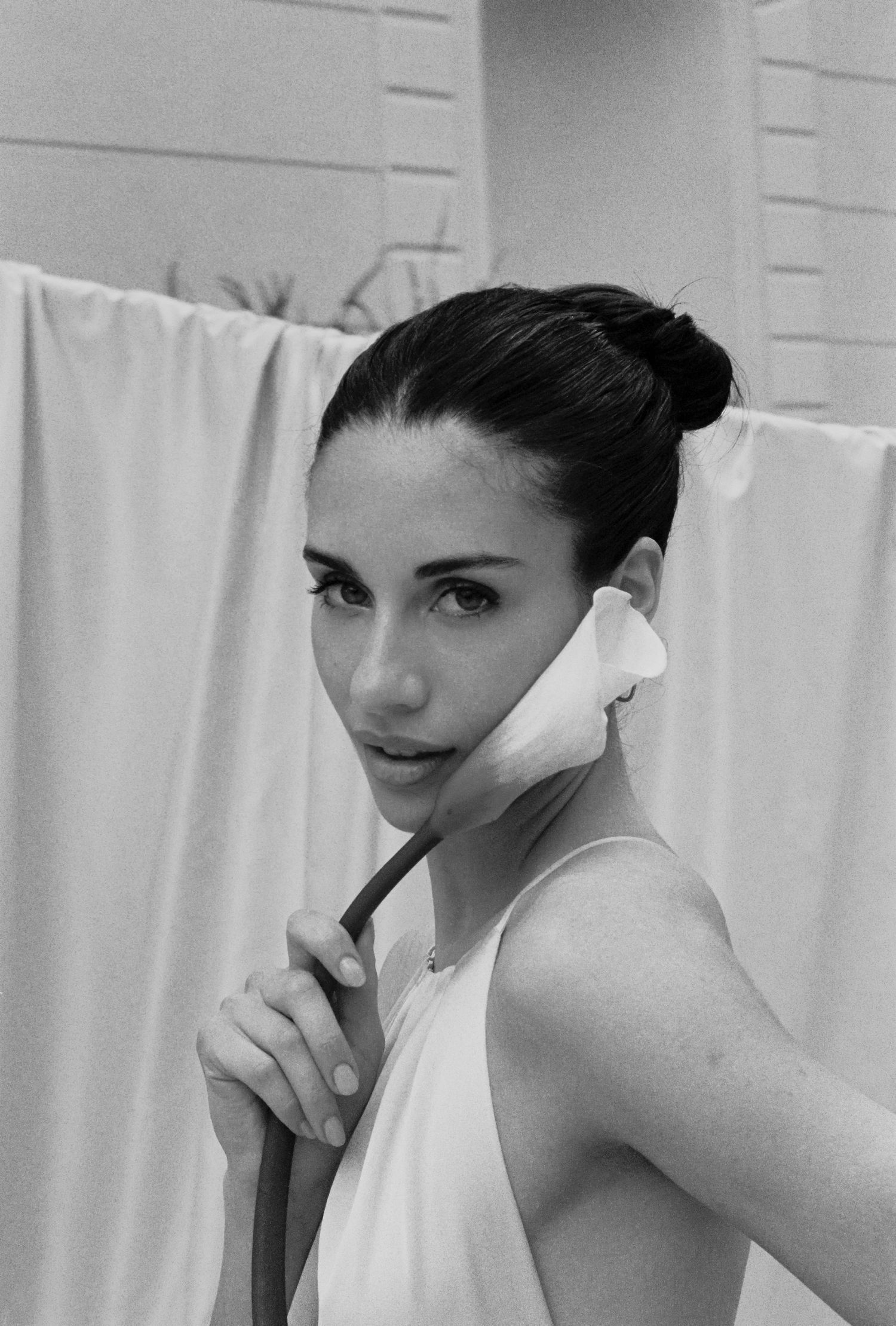 Black and white photo of a young woman holding a calla lily flower against her face, wearing a white dress.