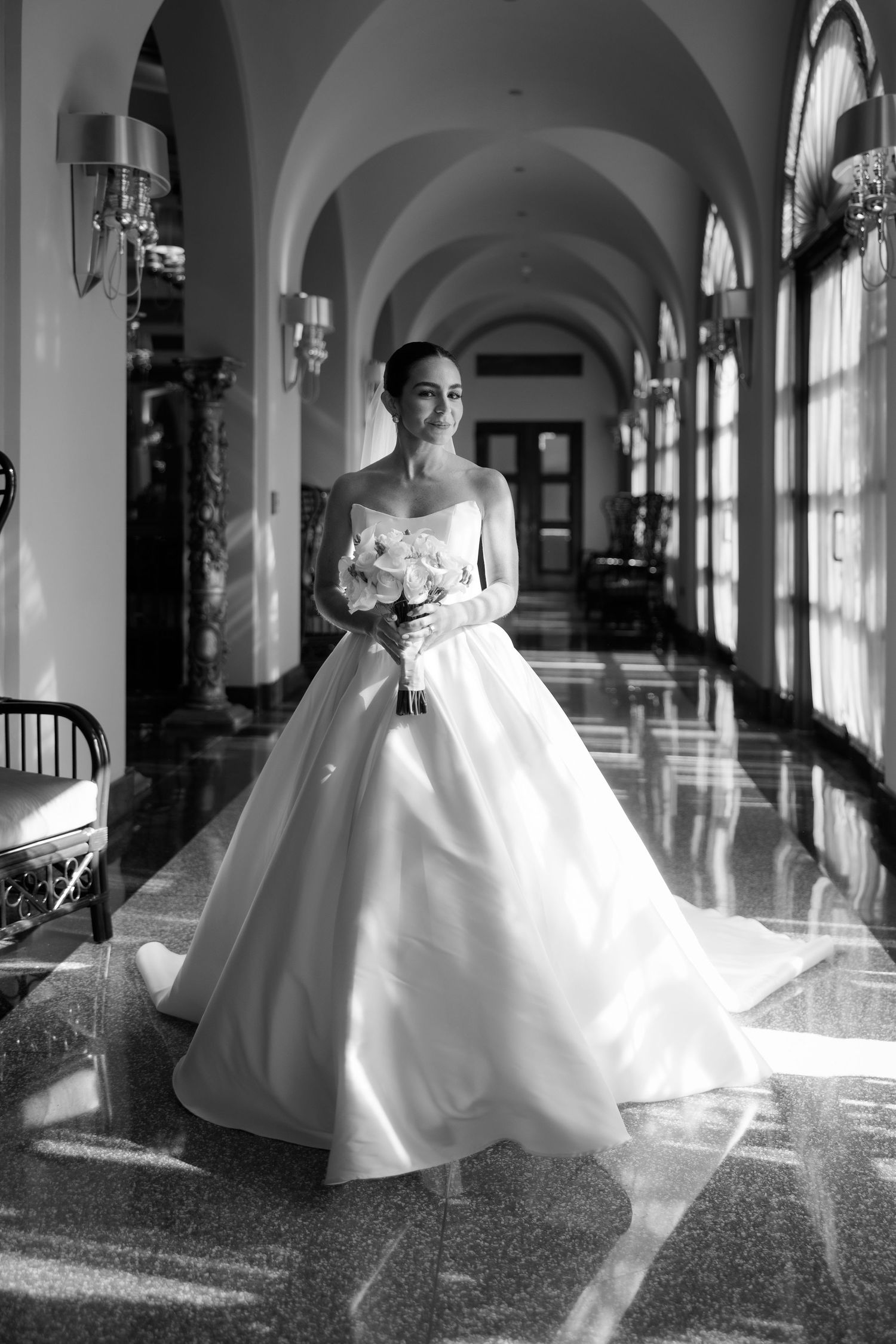Bride in a strapless ball gown holds bouquet in an elegant arched hallway, black and white wedding photo.