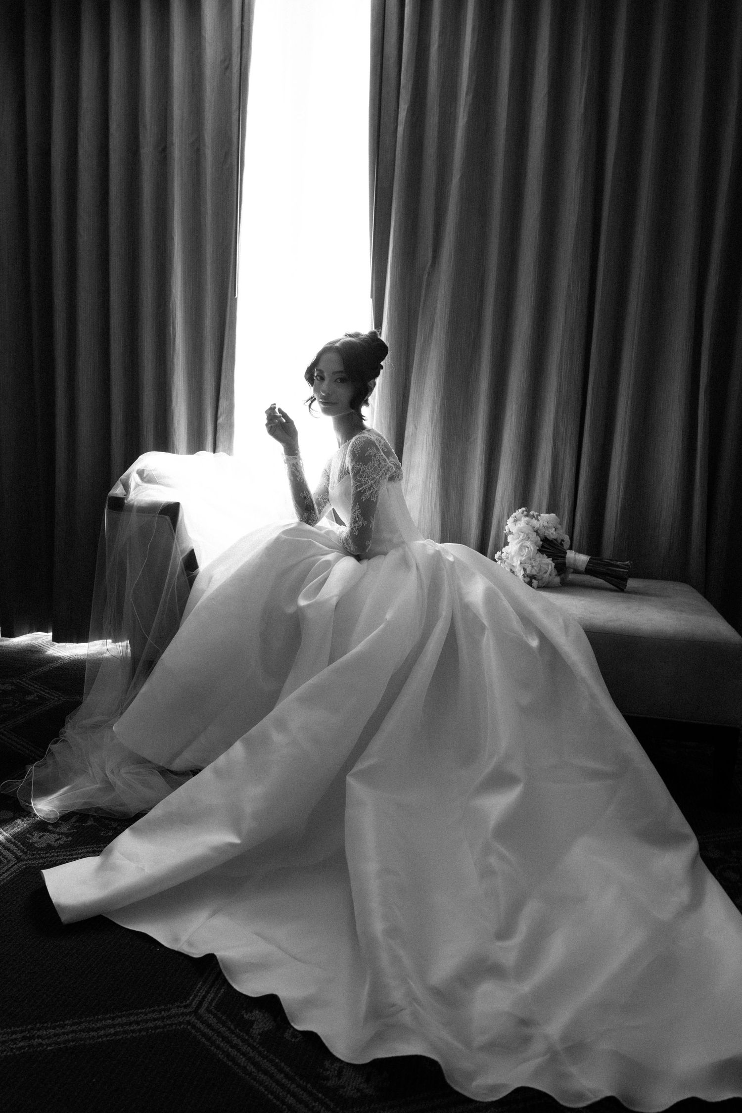 Black and white photo of a bride in a ballgown wedding dress sitting by a window with curtains.
