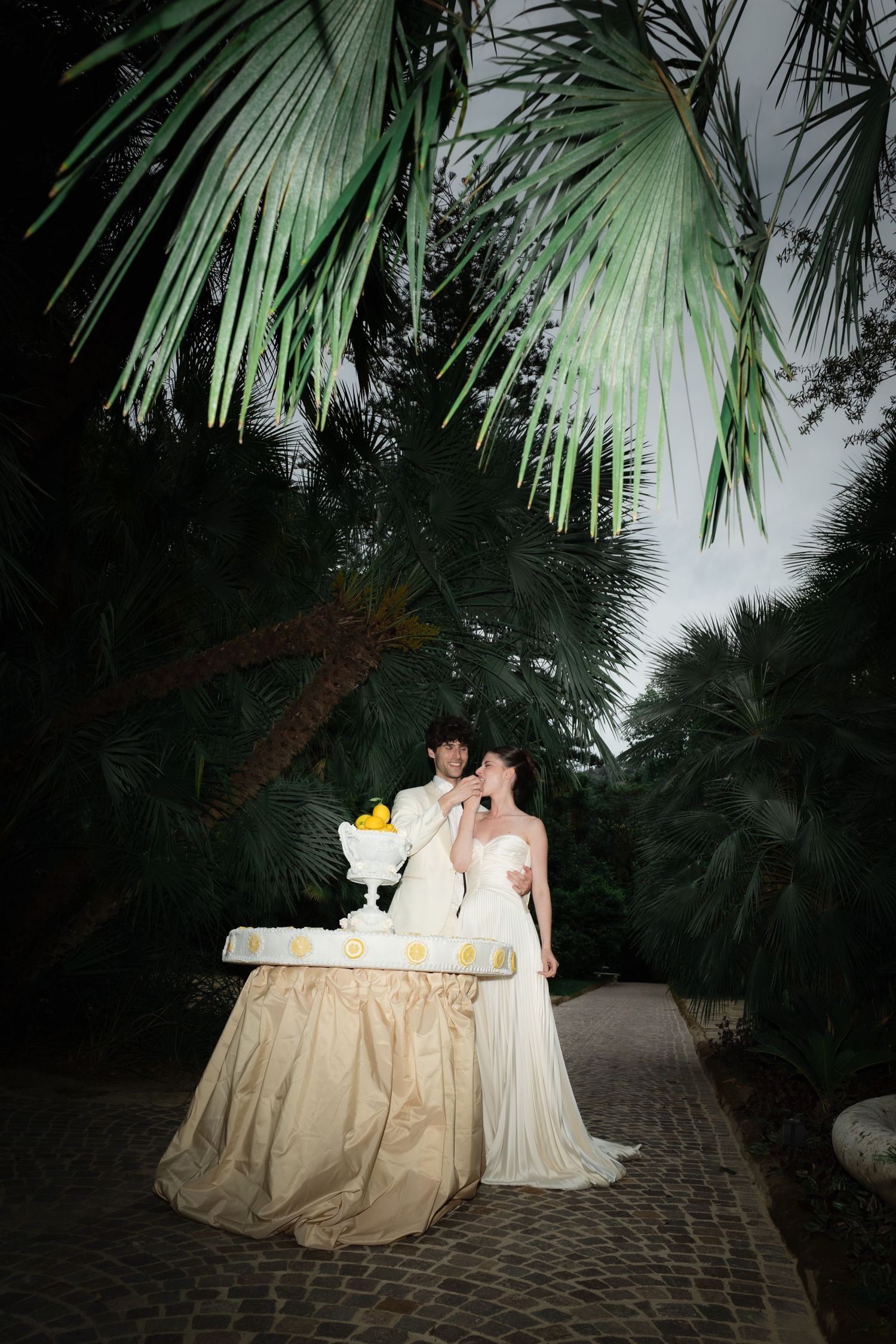 Bride and groom share a romantic moment beside their wedding cake outdoors at night, surrounded by lush palm trees.