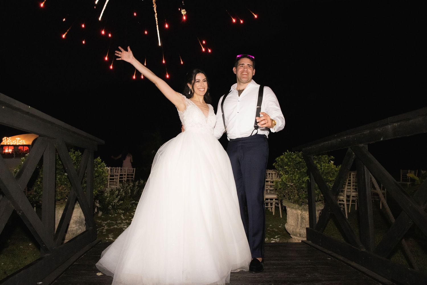 Newlyweds pose together on wooden bridge at night with bright lights in background.