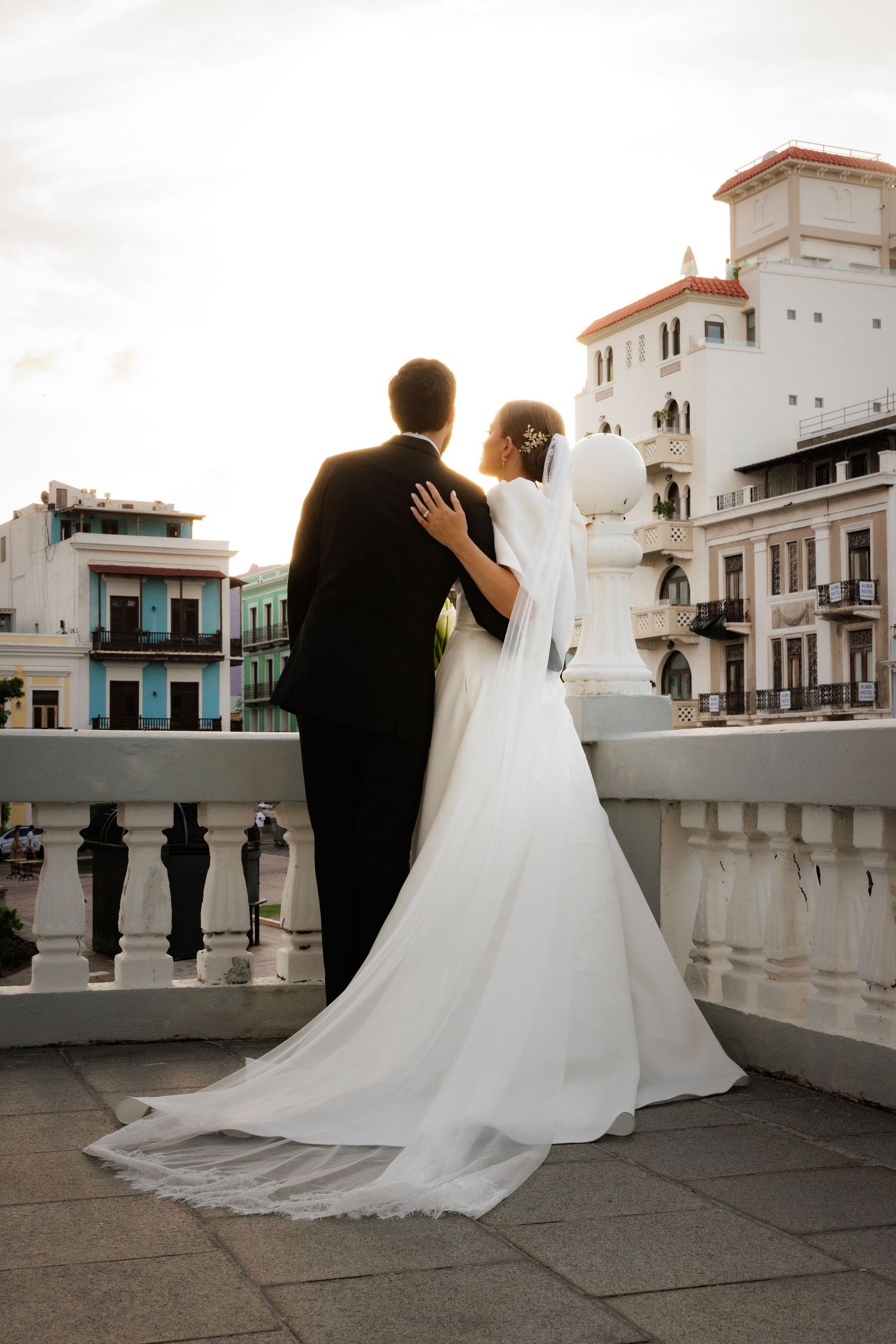 A bride and groom share an intimate moment on a balcony overlooking a historic building with white architecture at sunset.