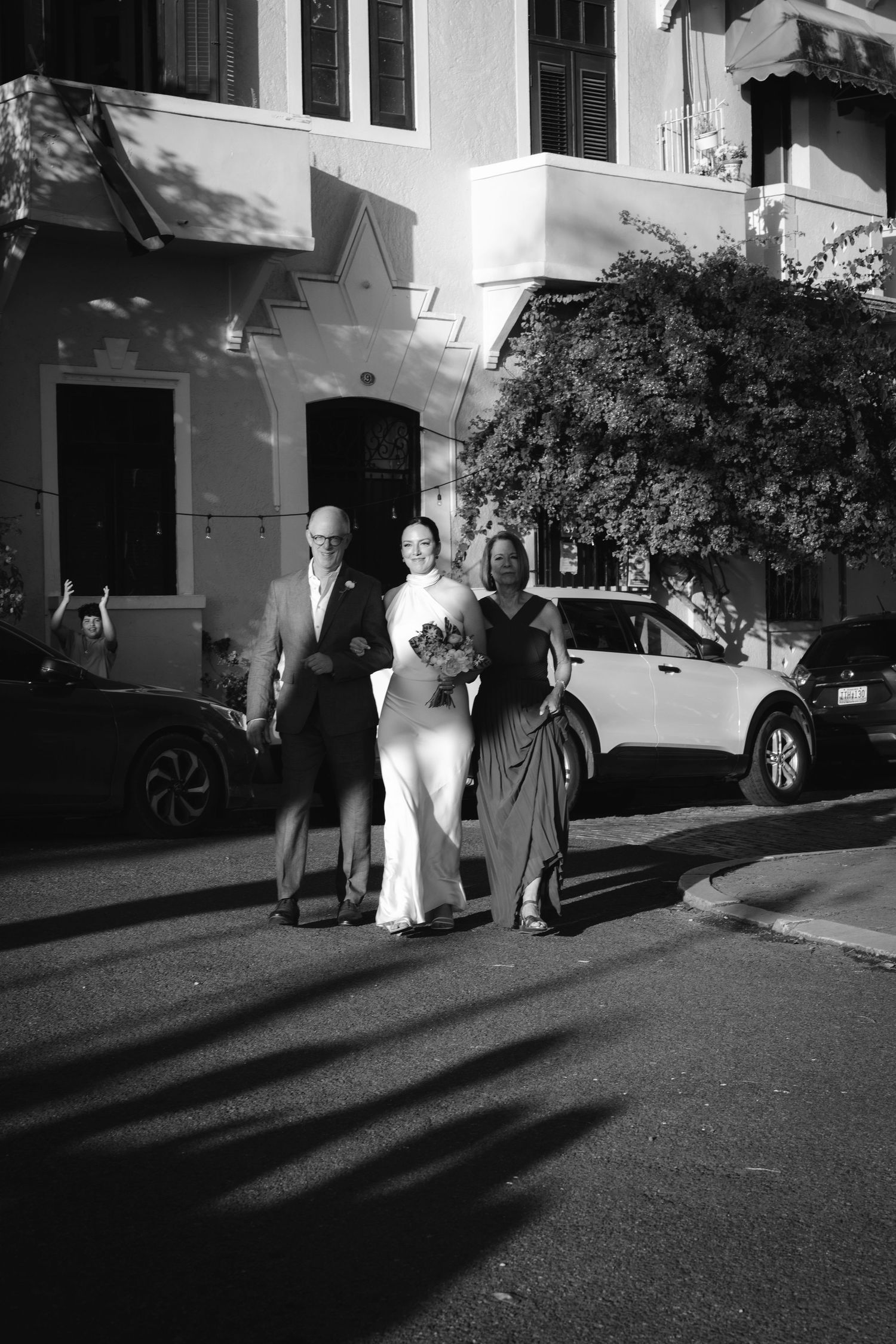 Black and white photograph of wedding guests walking along a sidewalk casting long dramatic shadows.