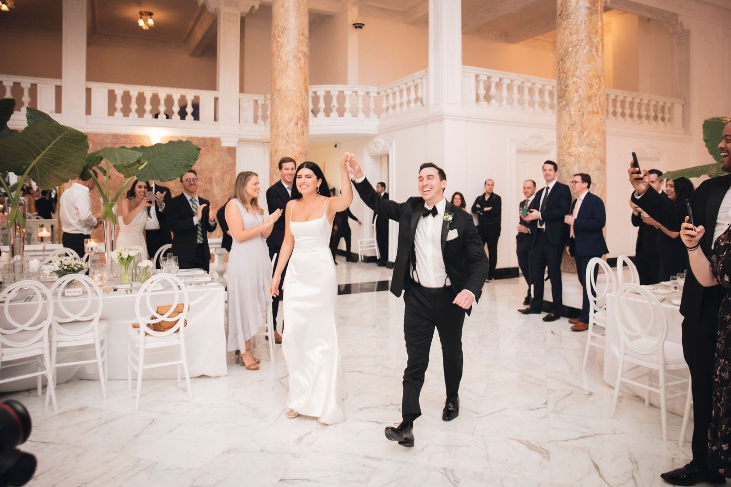 A celebratory wedding moment in an elegant marble columned ballroom with white chairs and guests looking on.