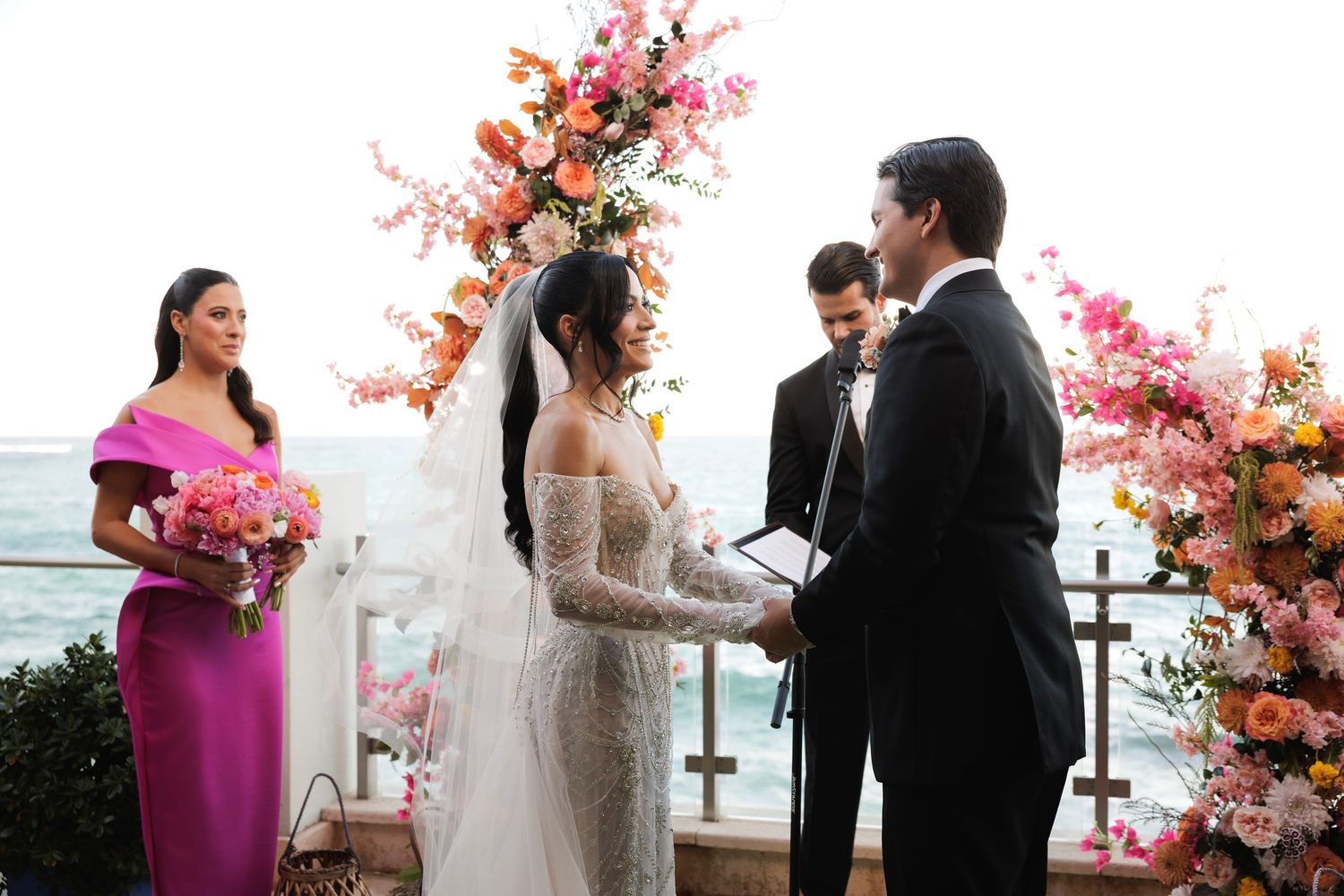 A waterfront wedding ceremony features guests seated under pink cherry blossom floral arrangements by the ocean.