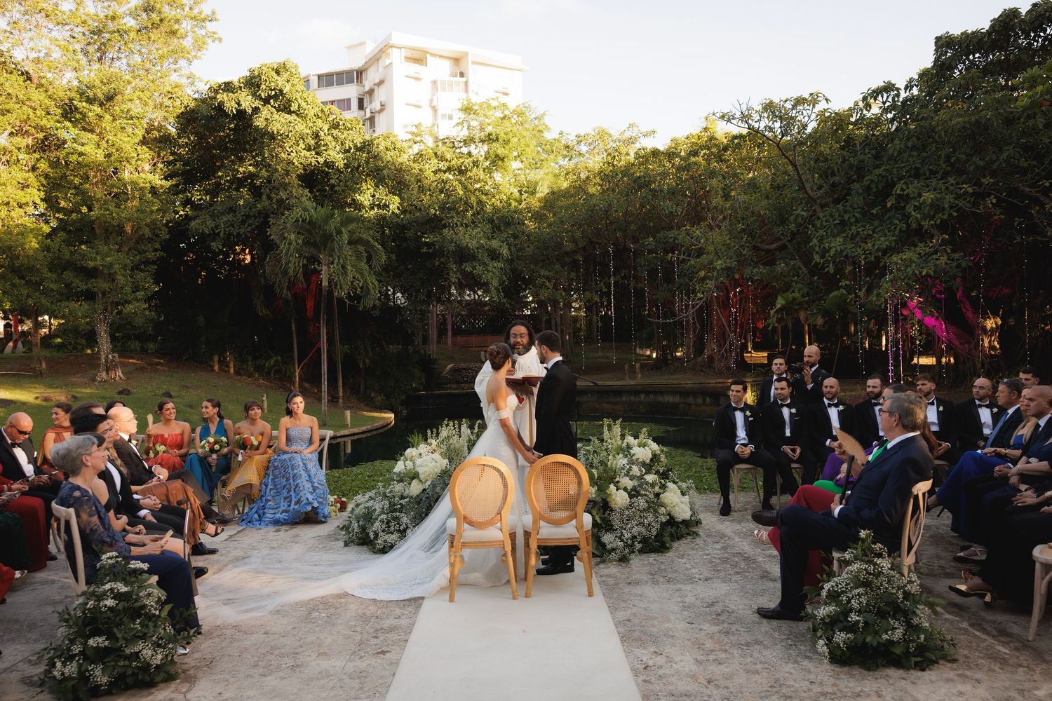 A couple shares their wedding ceremony in a picturesque outdoor garden setting with guests seated on both sides of white aisle.