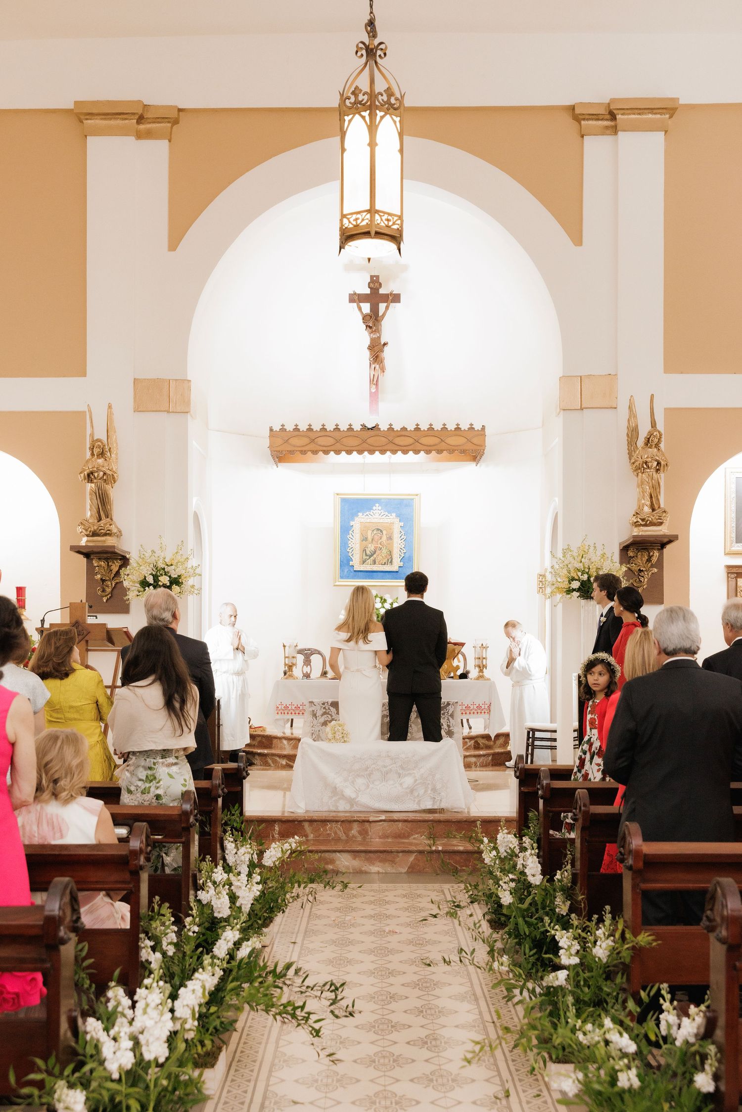 A church interior decorated with white floral arrangements during a wedding ceremony with people in pews and an arched altar.