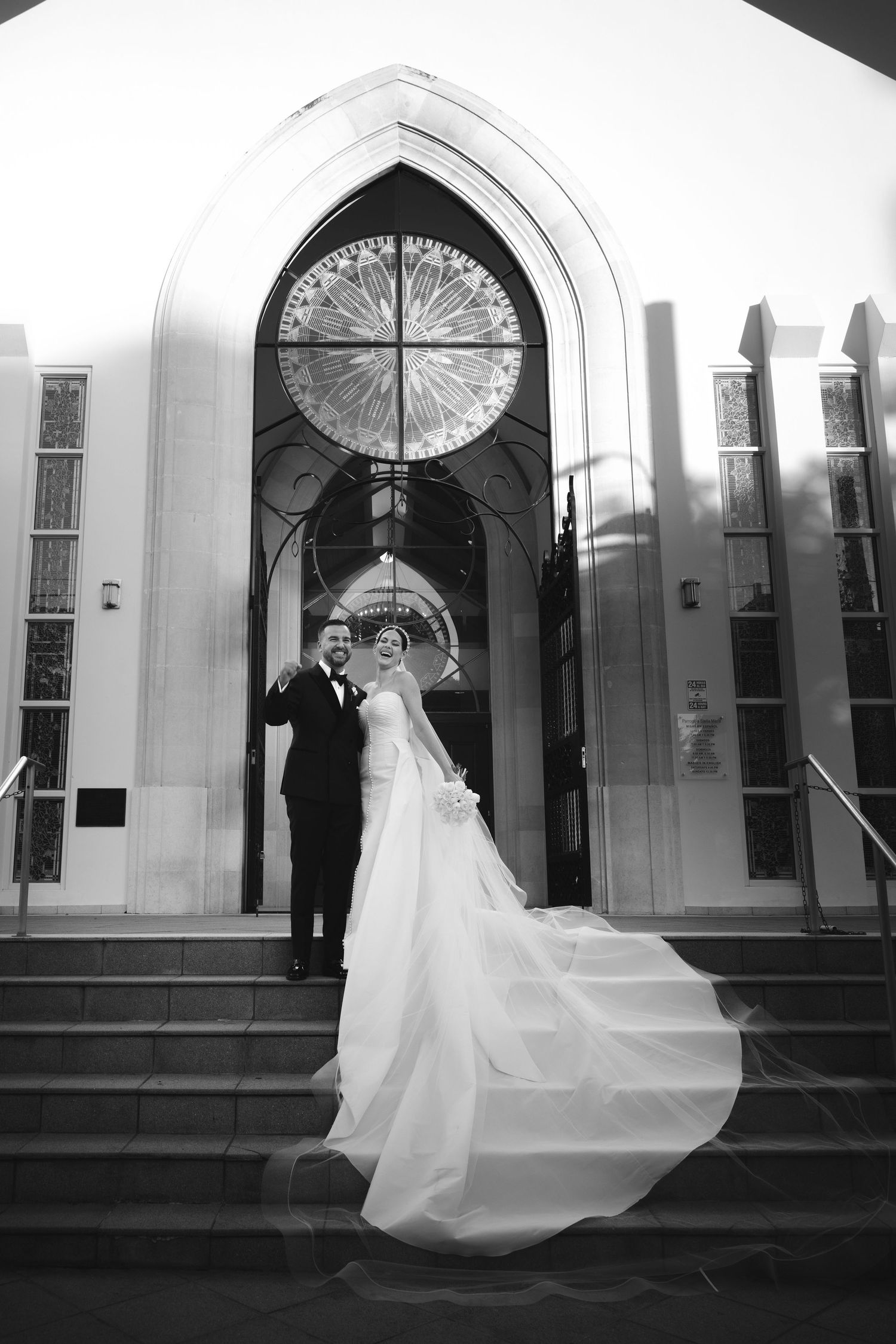 A wedding couple stands in front of a church entrance with an arched doorway and circular stained glass window above.