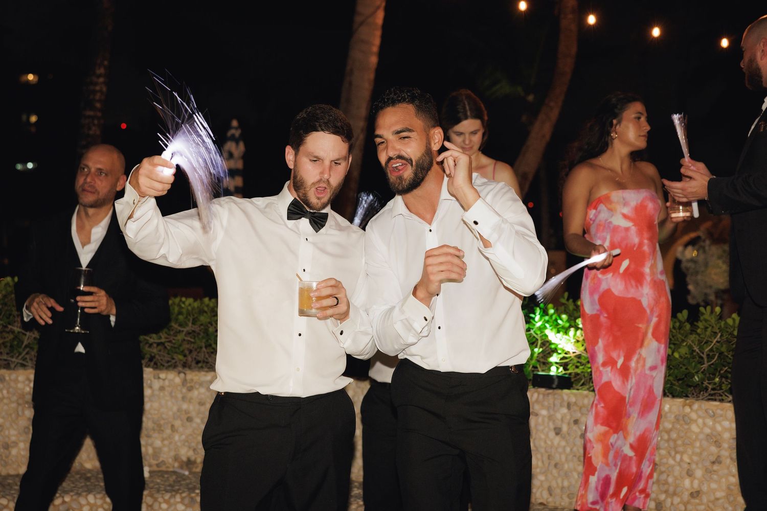 Wedding guests in formal attire celebrate at an outdoor evening reception with string lights and palm trees.