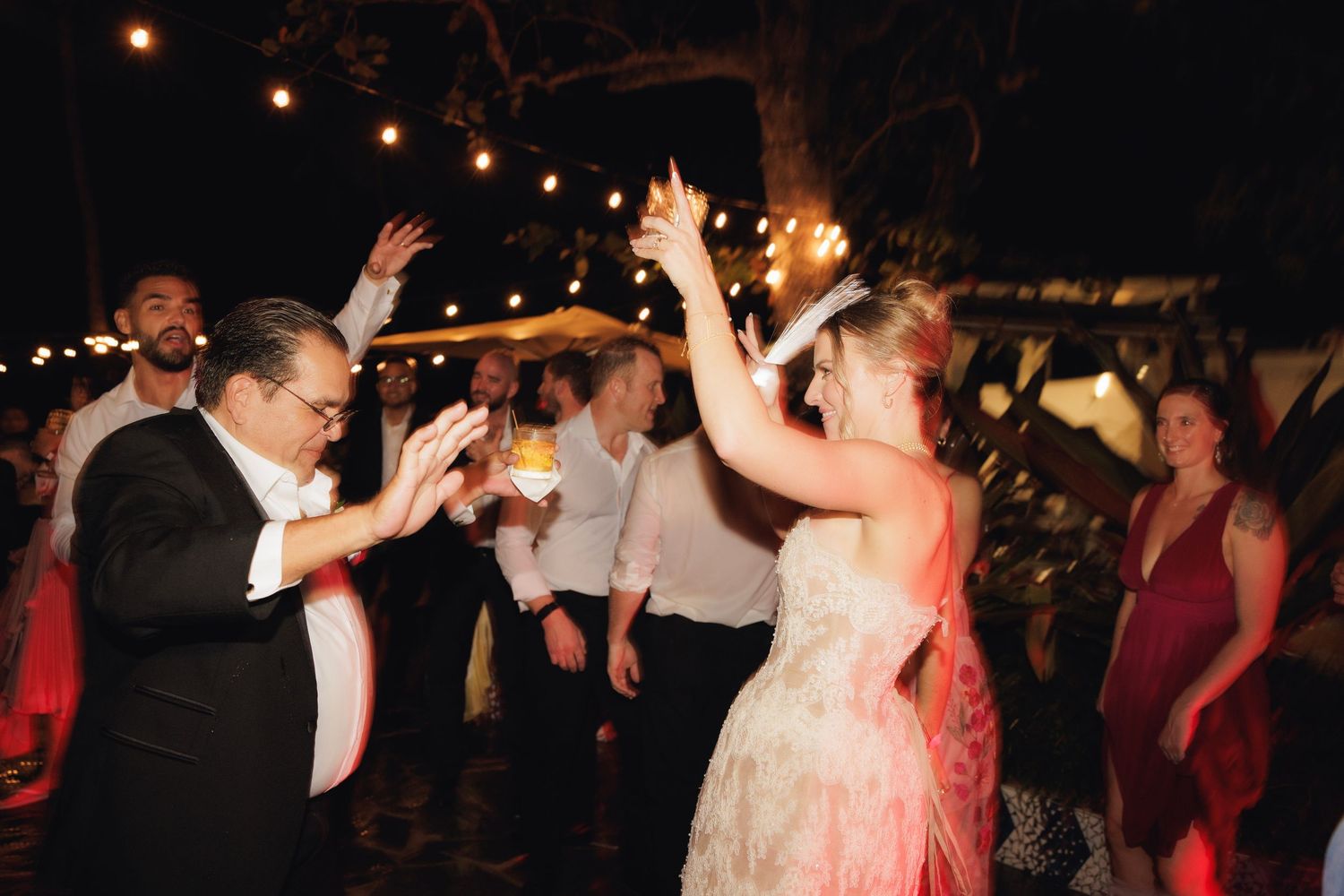 People dancing together at an outdoor evening wedding reception with string lights overhead and a lively party atmosphere.