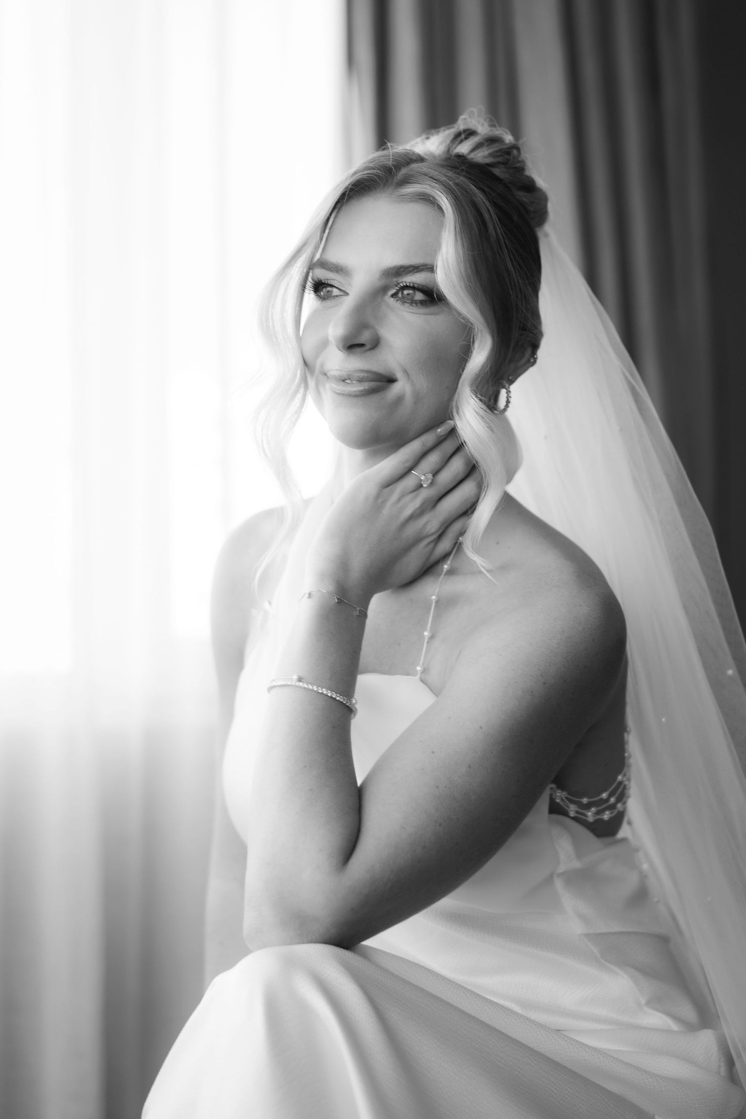 A black and white portrait of a bride in a wedding dress and veil, touching her neck softly while sitting and smiling.