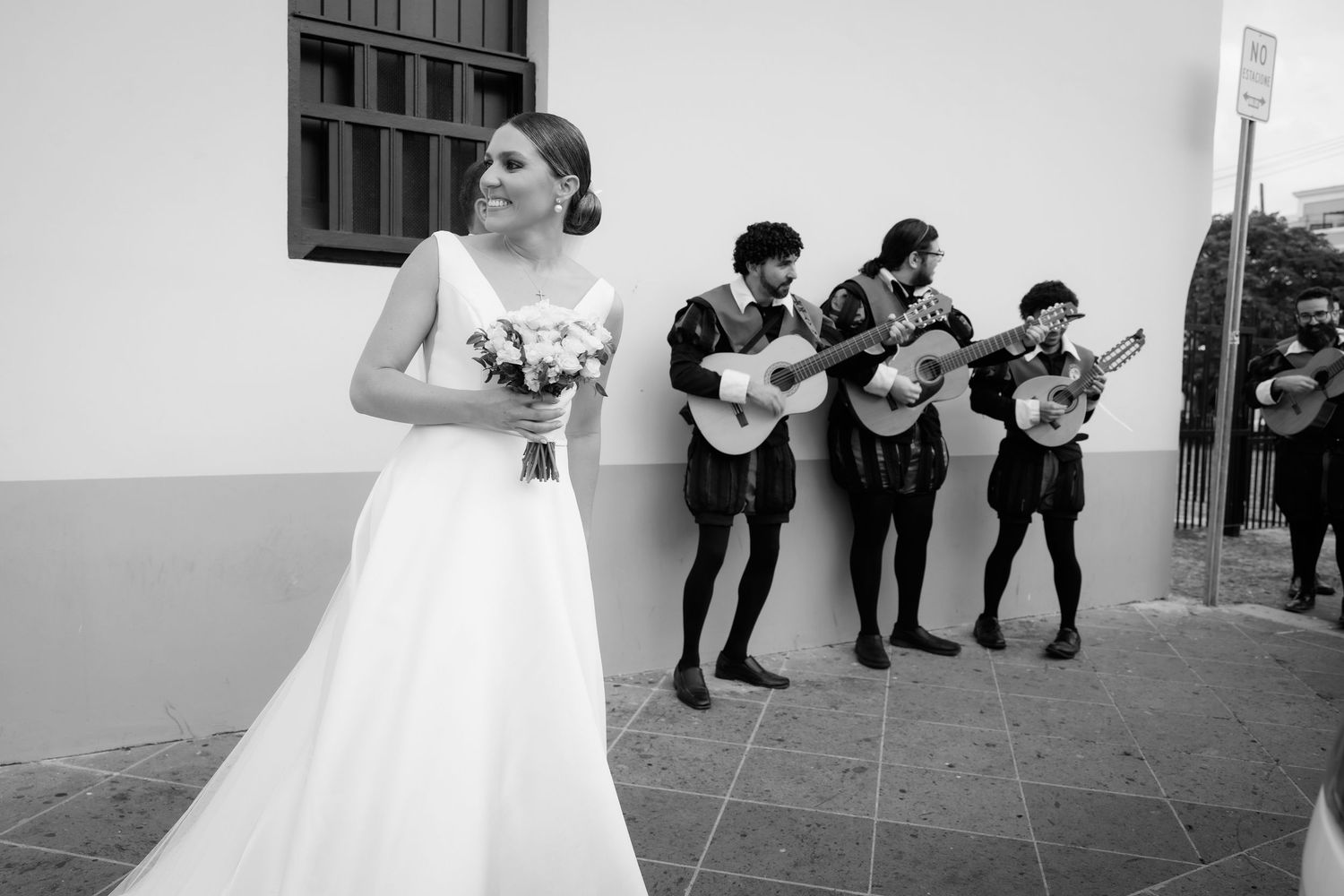 Bride in white dress stands with bouquet while mariachi band performs in background.