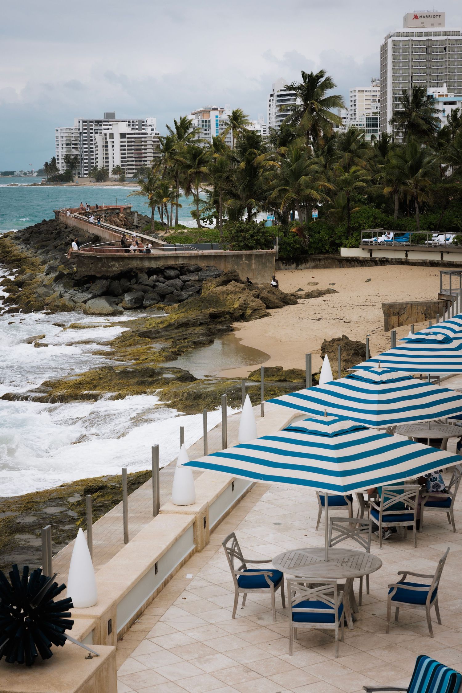 Beachfront patio with blue and white striped umbrellas overlooking rocky coastline and high-rise buildings in Puerto Rico.