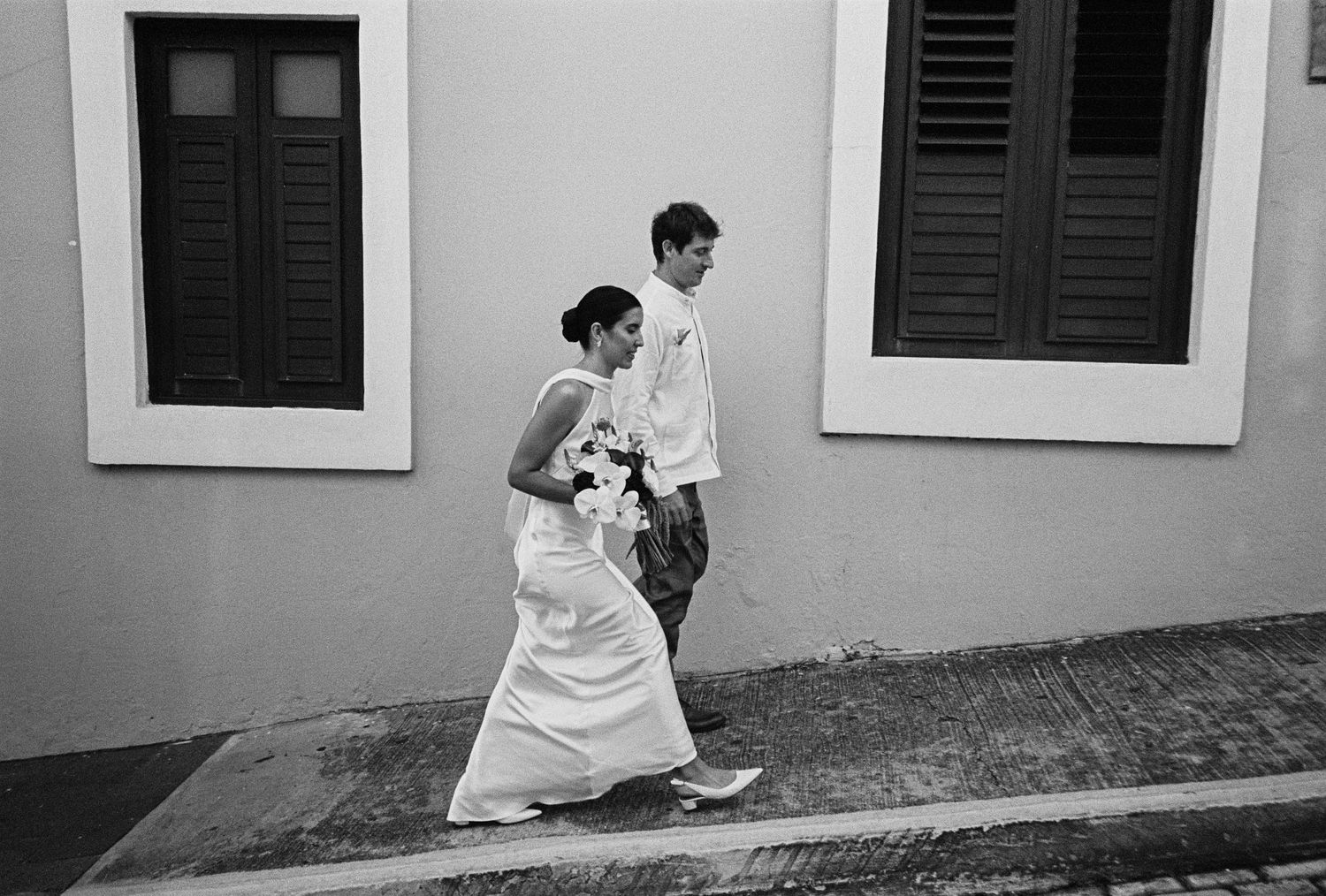 Black and white photo of a bride in white gown and groom walking along a cobblestone street past a pastel building.