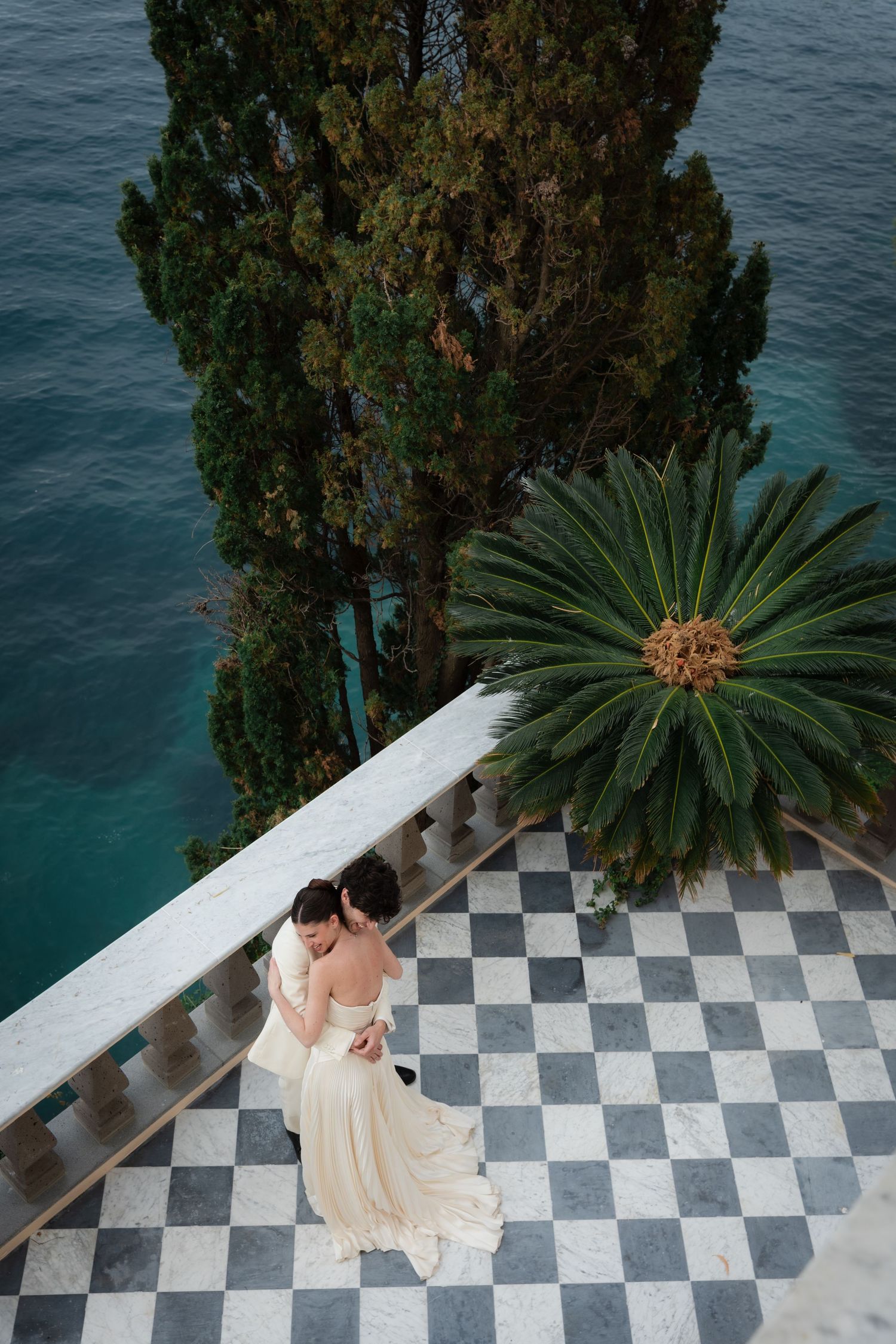 Bride and groom embrace on a checkered marble terrace overlooking turquoise sea, with cypress and palm trees nearby.