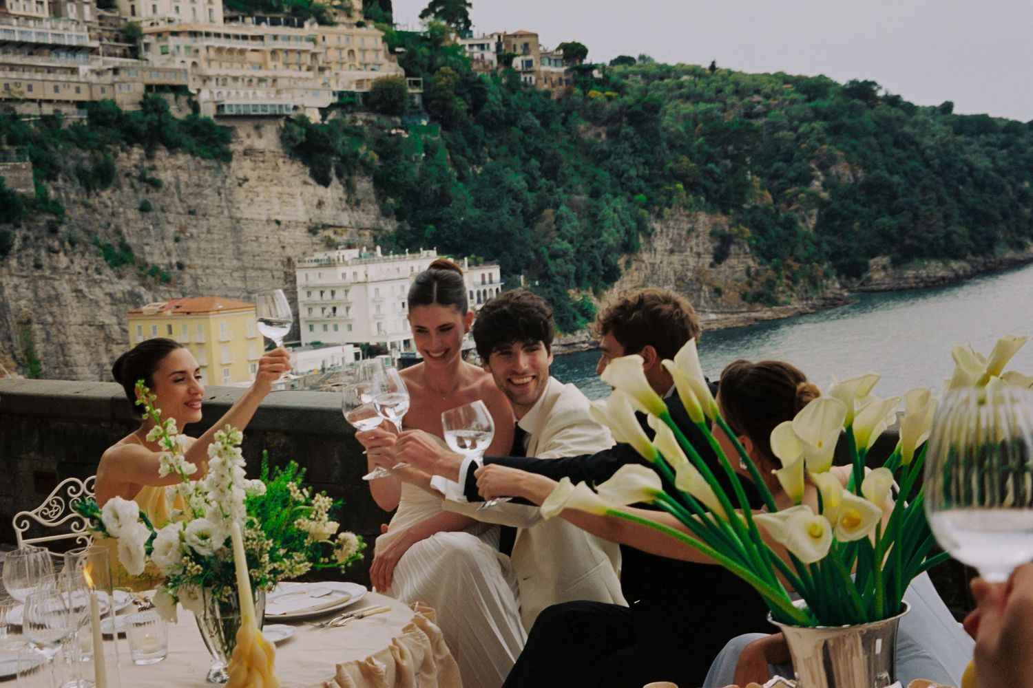 Elegant outdoor dinner party on an Amalfi Coast terrace with guests toasting wine glasses surrounded by white calla lilies.