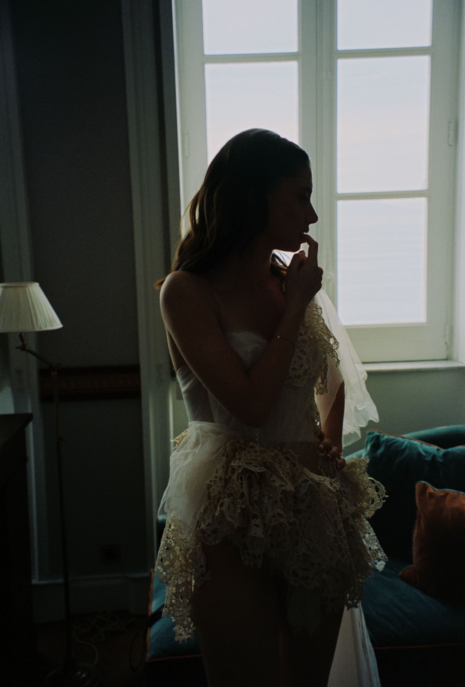 Bride in lace wedding dress and veil standing by window in soft backlight before ceremony.