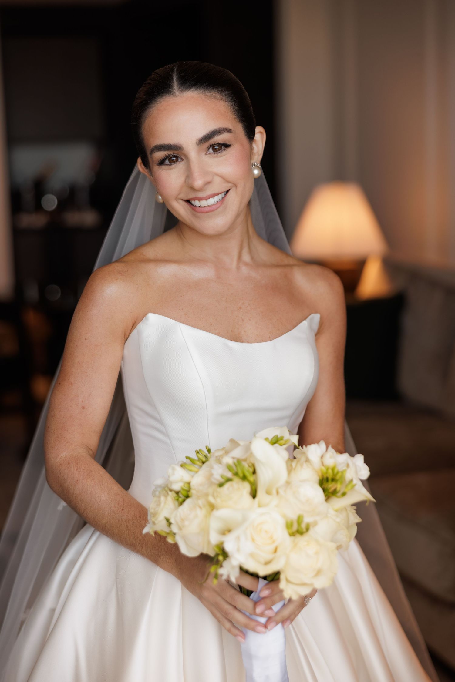 A bride in a strapless white wedding gown holds a bouquet of cream roses and greenery.