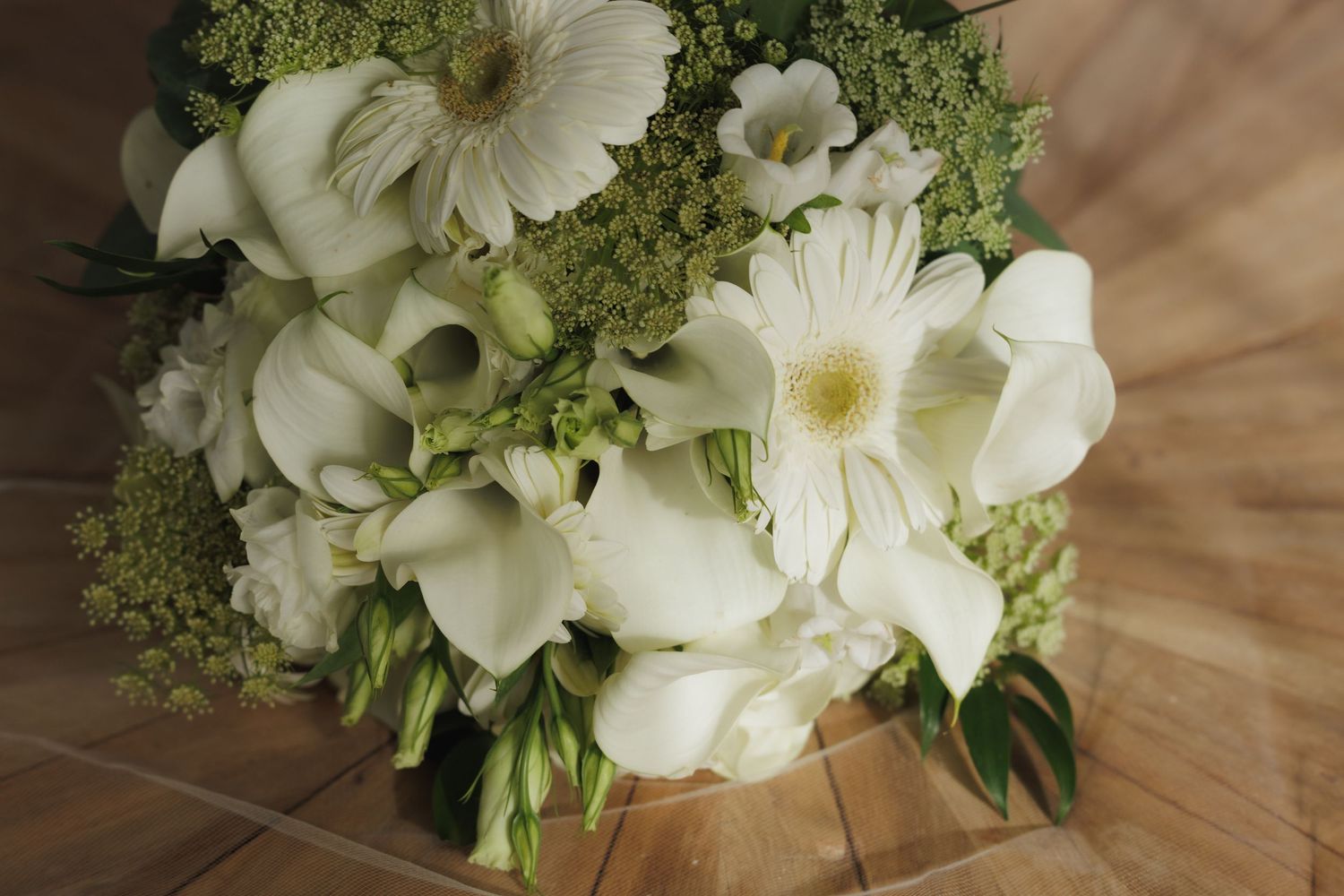 White gerbera daisies and delicate greenery arranged in an elegant wedding bouquet on wooden surface.