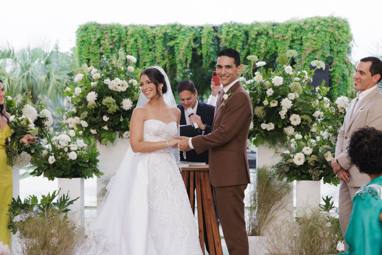 Wedding ceremony scene with lush greenery and white rose arrangements framing the moment.
