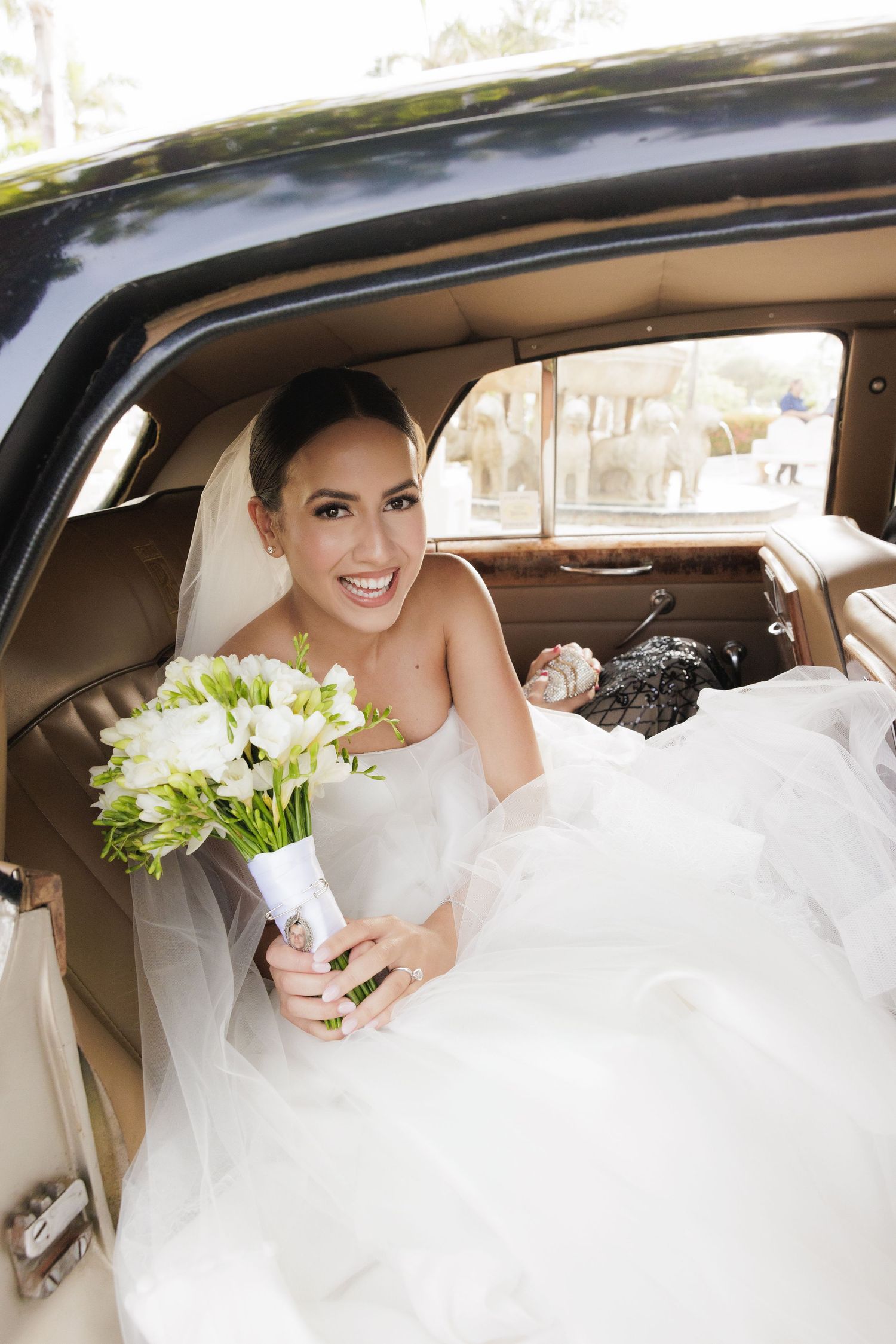 A wedding scene showing a bride in a white dress sitting in a luxury car with a bouquet of white flowers.