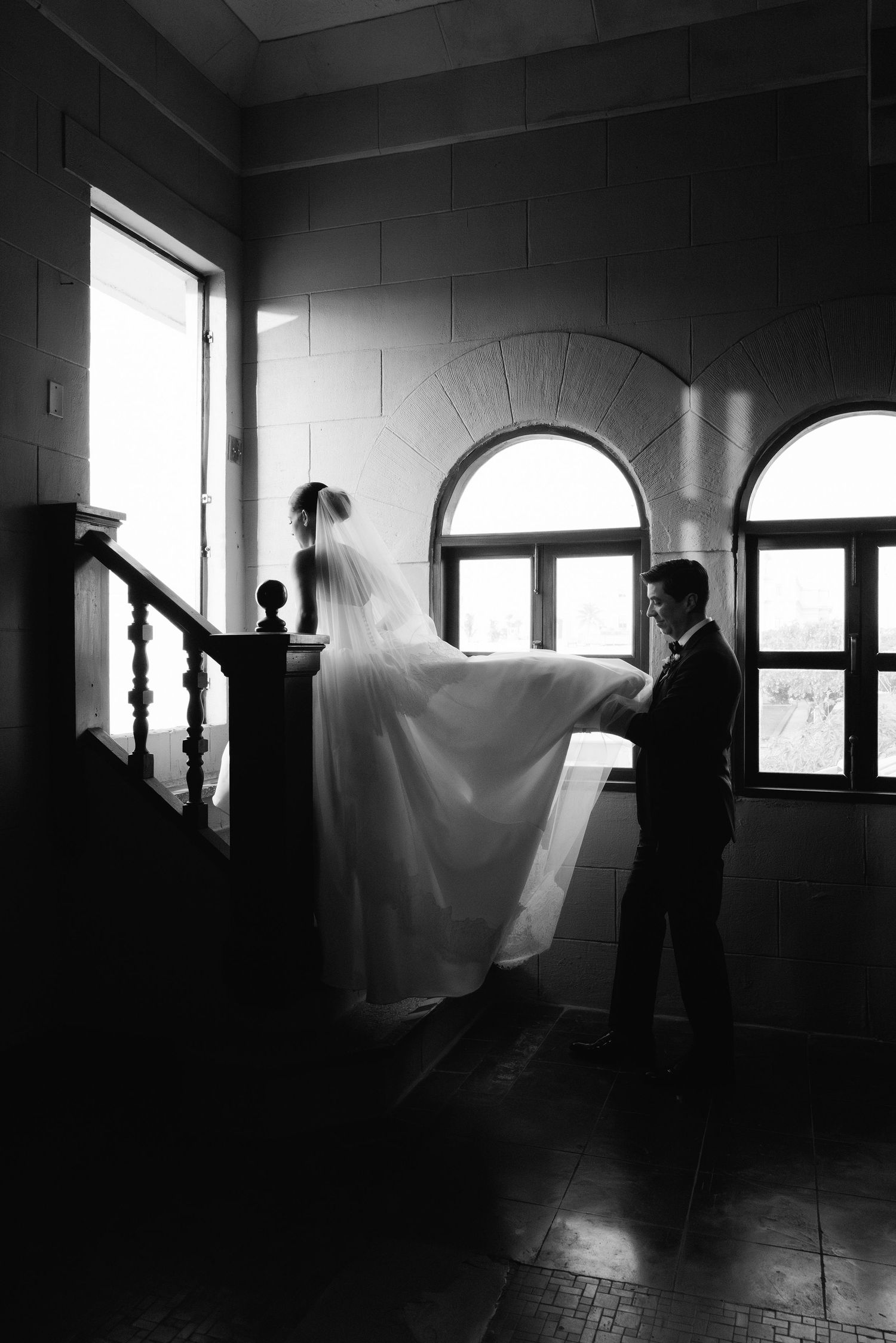 A dramatic black and white wedding photo showing silhouettes against arched windows in an elegant stairwell.