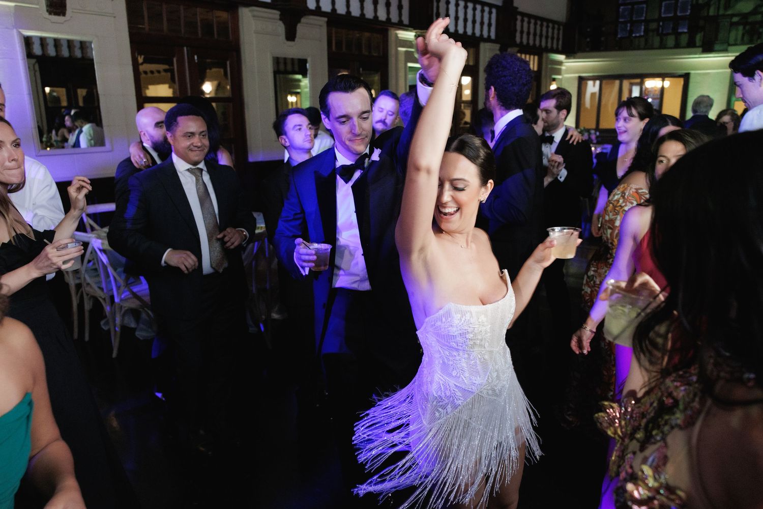 Wedding guests dance and celebrate on a lively dance floor illuminated by purple lighting at an evening reception.