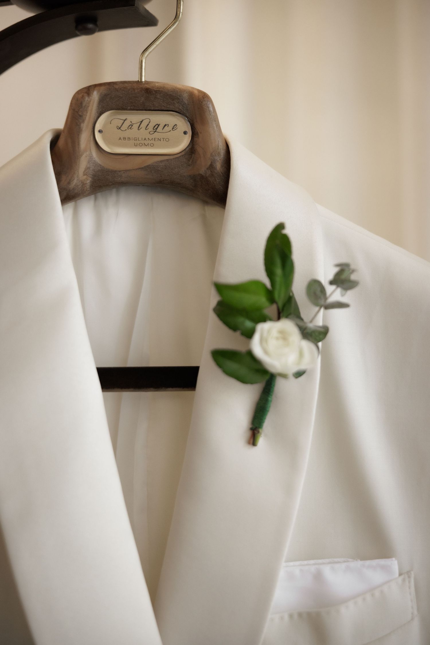 A white boutonniere with delicate flowers and green leaves is pinned to a formal white suit lapel.