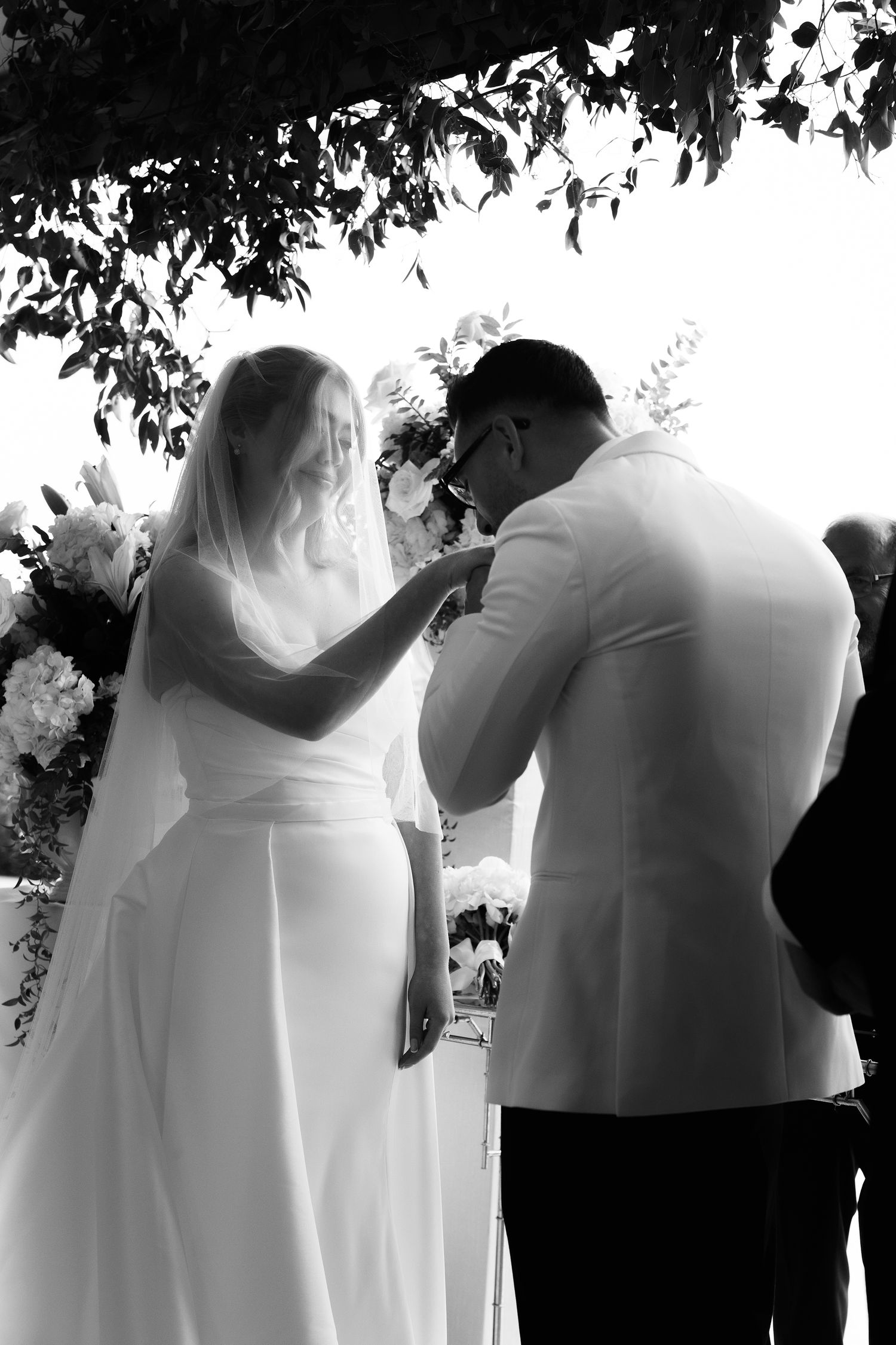 Black and white photo of a tender moment during an outdoor wedding ceremony under tree branches.