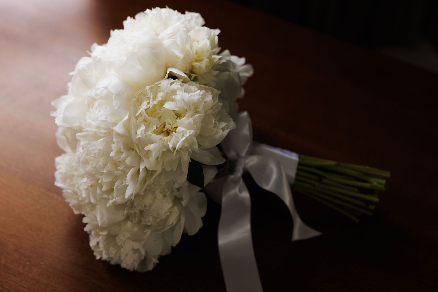 White peony bouquet with satin ribbon rests on dark wooden surface in elegant wedding styling.