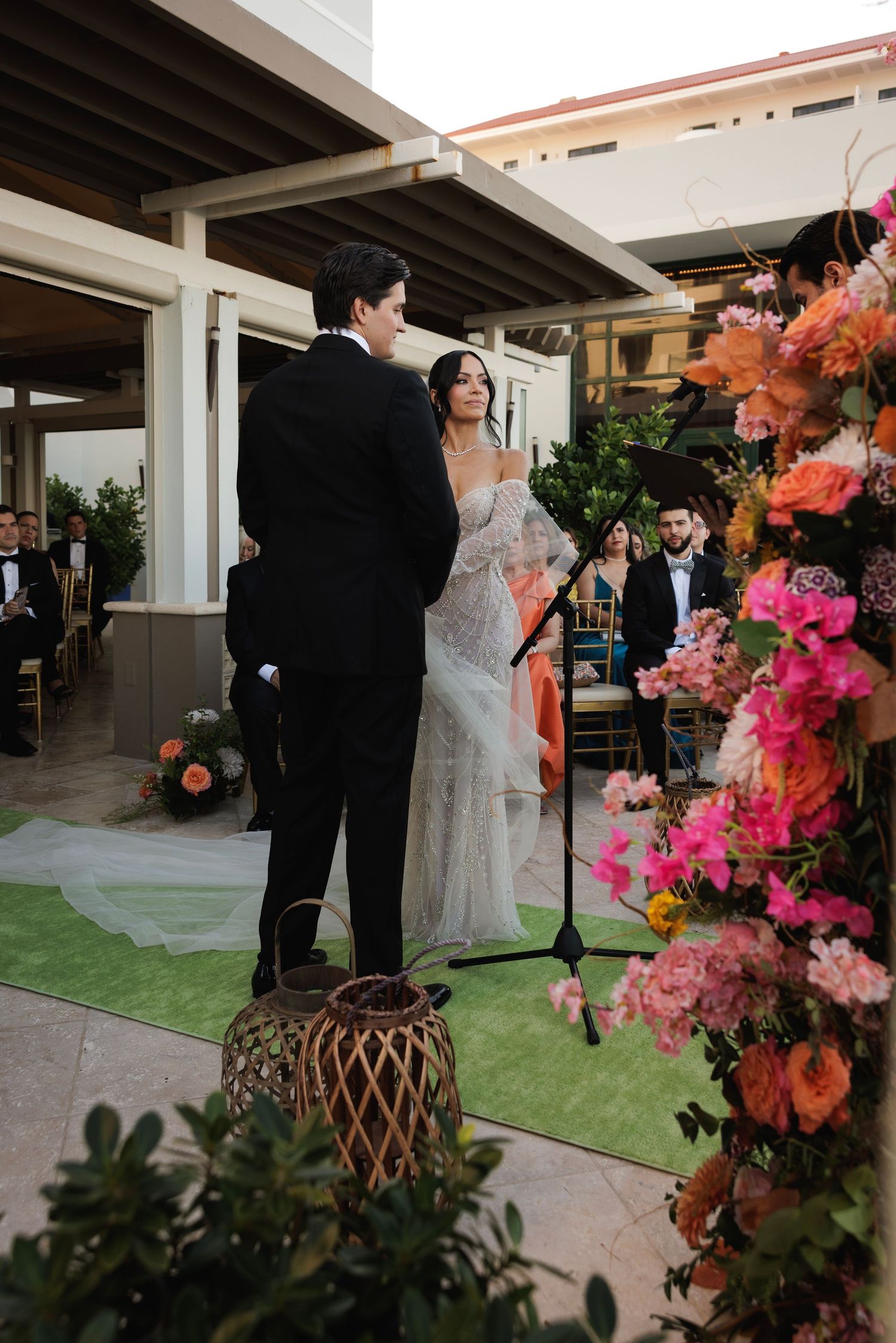 Wedding ceremony at outdoor venue with green carpet aisle runner and vibrant pink and orange floral arrangements.