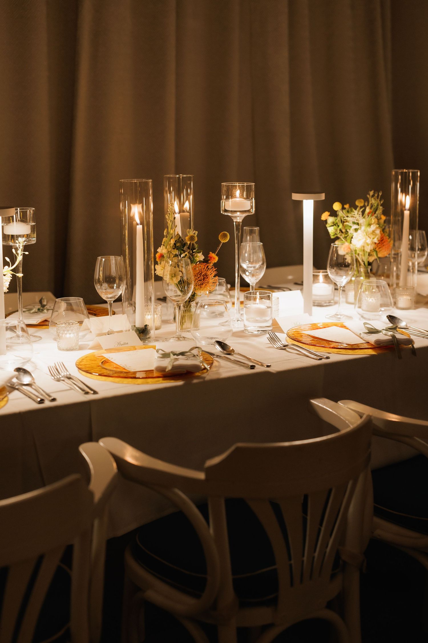 An elegant wedding reception table setting with white linens, glowing candles, and floral centerpieces in a dimly lit room.