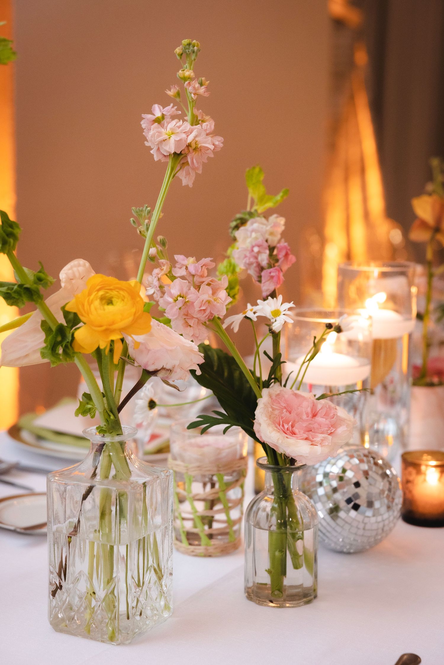 Simple wedding centerpieces with delicate pink and yellow flowers in glass jars alongside mercury glass votives.