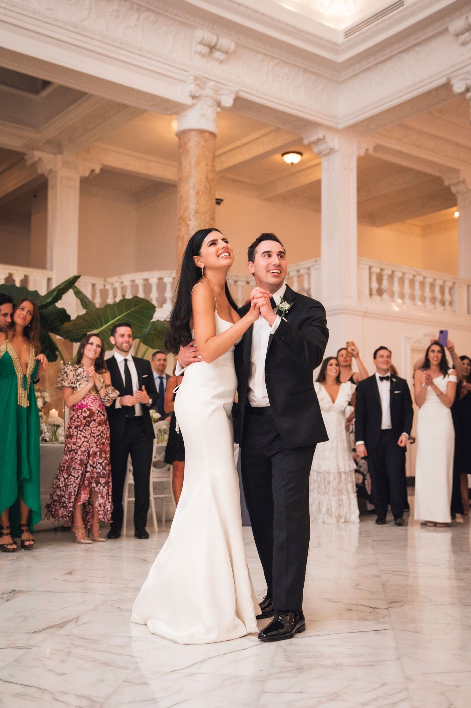 A couple shares their first dance in a grand ballroom with marble columns and ornate ceiling details.