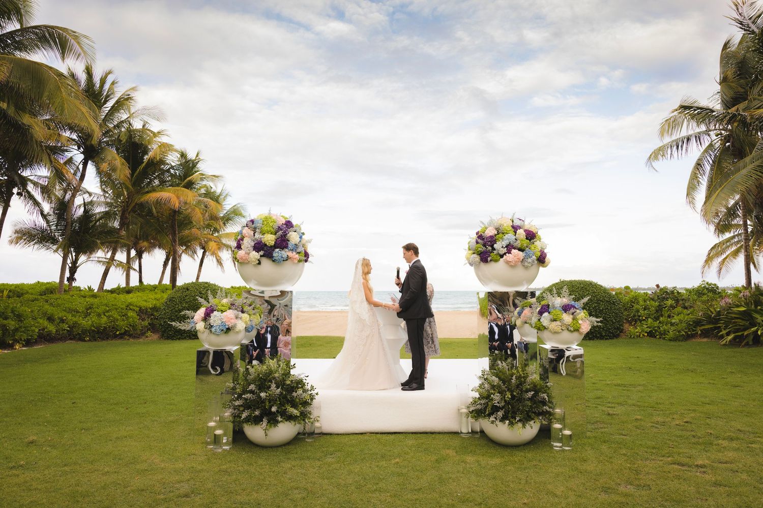 A tropical beach wedding ceremony by the ocean with palm trees and white decor on a sunny day.