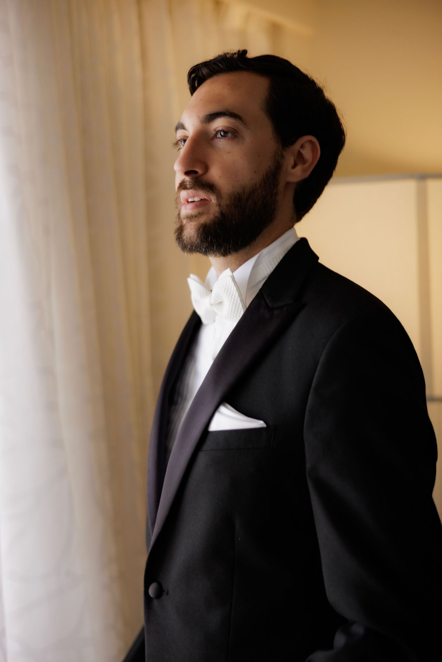 A groom in a classic black tuxedo and white bow tie gazes thoughtfully by a window.