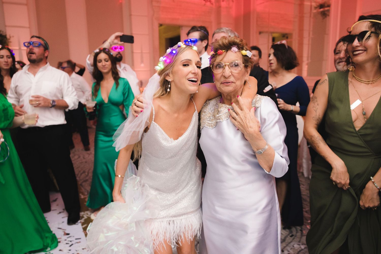 Wedding guests dance and celebrate at a pink-lit reception venue with people wearing white and green formal attire.