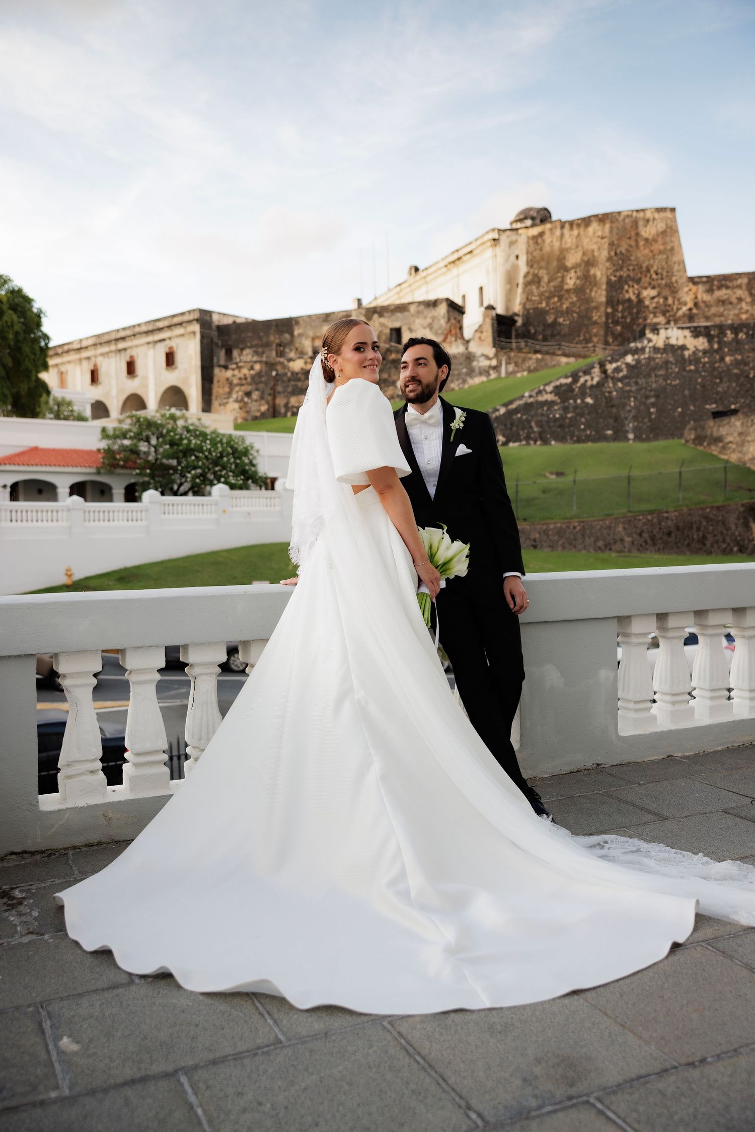 Newlyweds share an intimate moment on a stone balcony with a historic fortress wall backdrop.