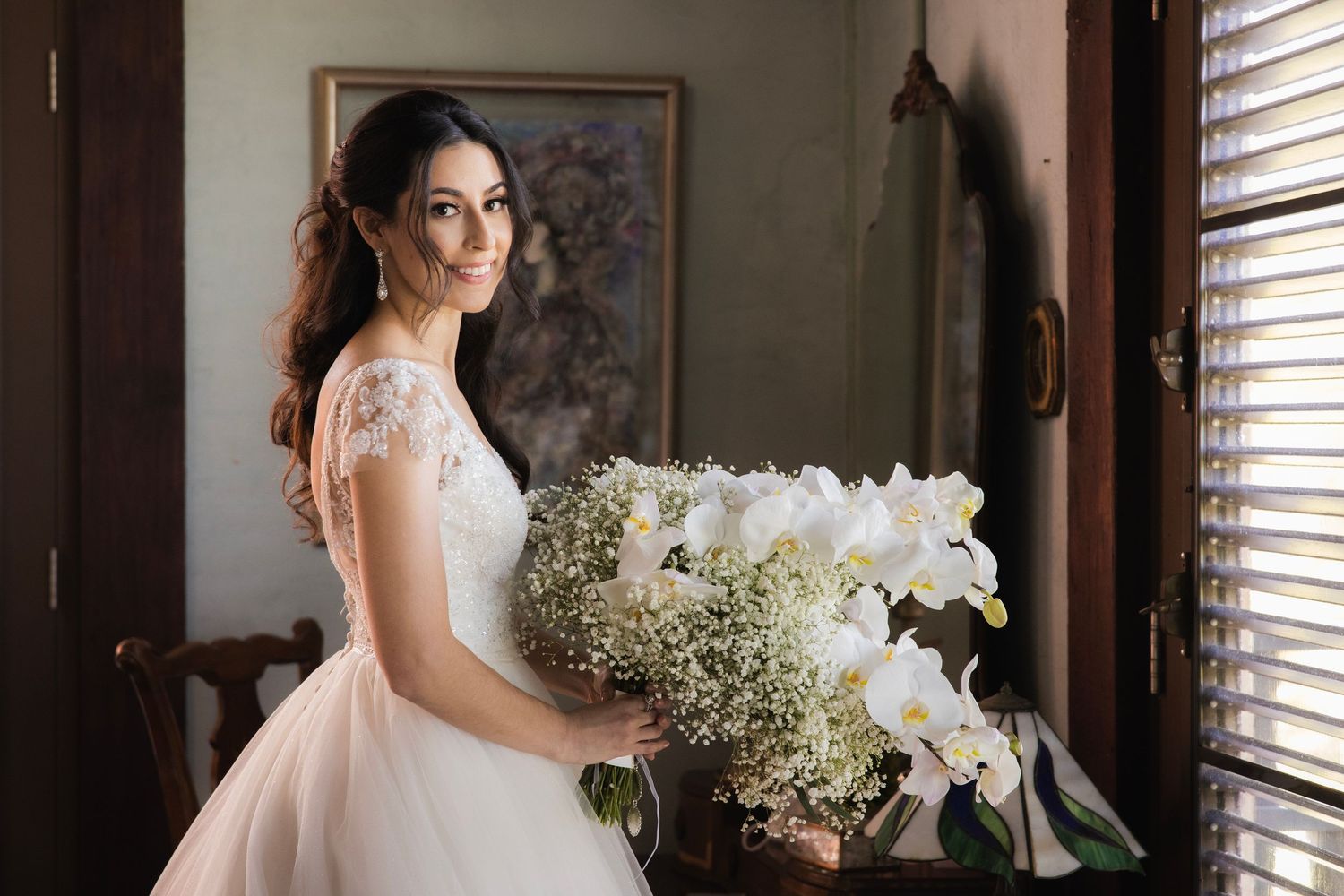 A bride in a classic white wedding gown sits near a window holding a bouquet of white orchids and baby's breath.