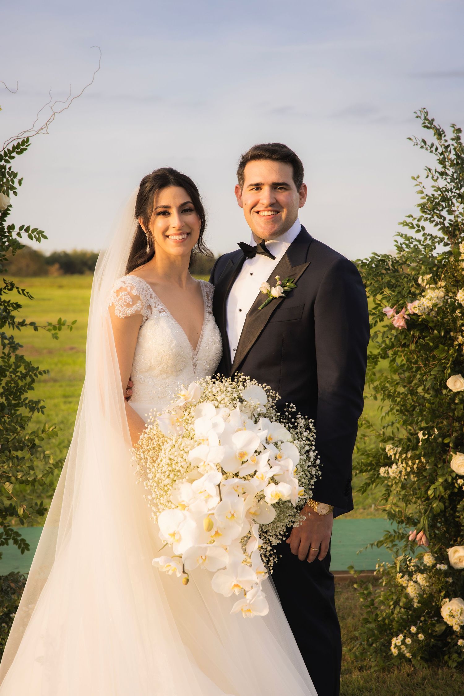 Elegant wedding couple posing in garden setting with white floral bouquet at sunset.