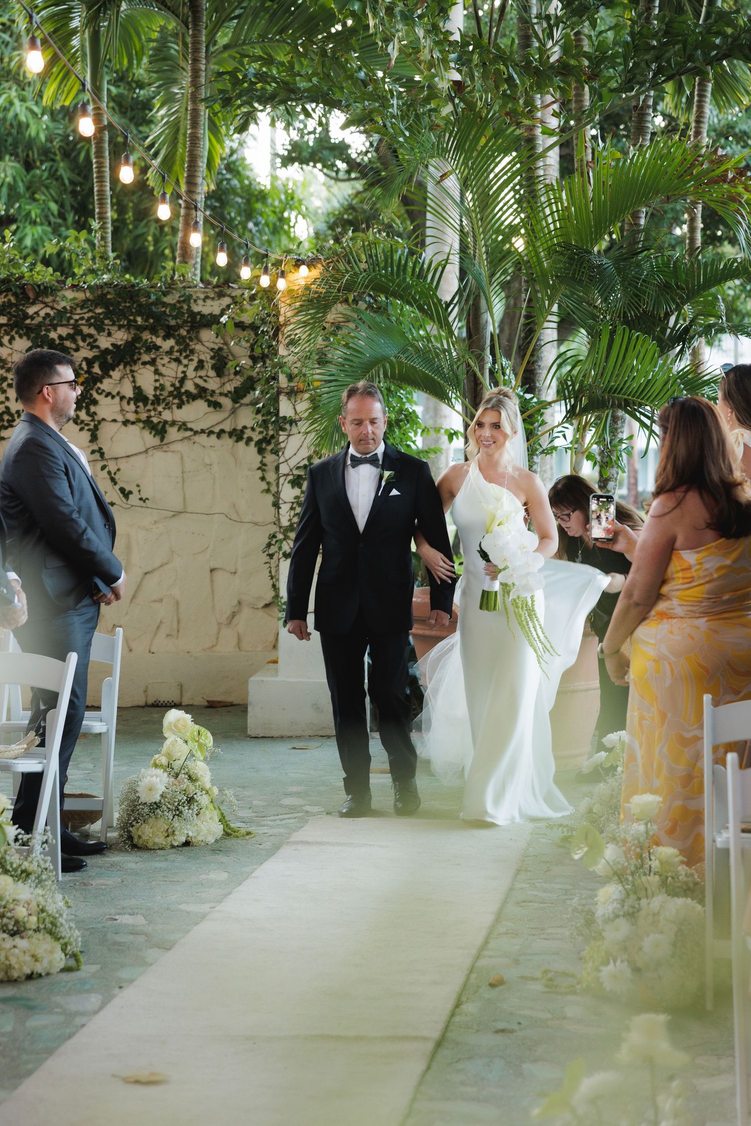 Elegant wedding ceremony taking place in a lush tropical venue with string lights and palm trees overhead.