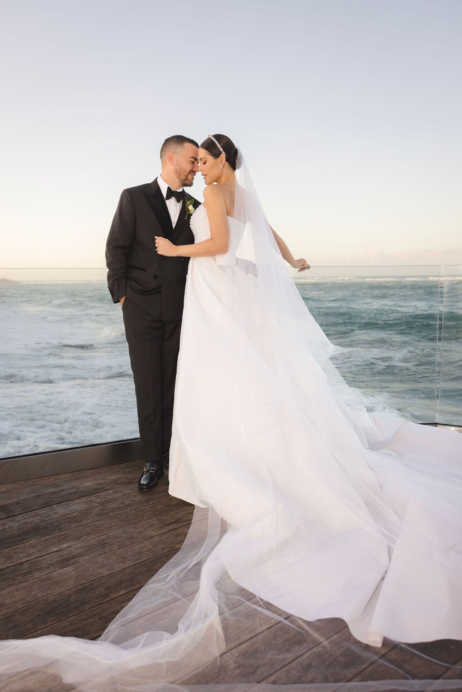 A bride and groom share an intimate moment on a wooden deck overlooking the ocean at sunset.