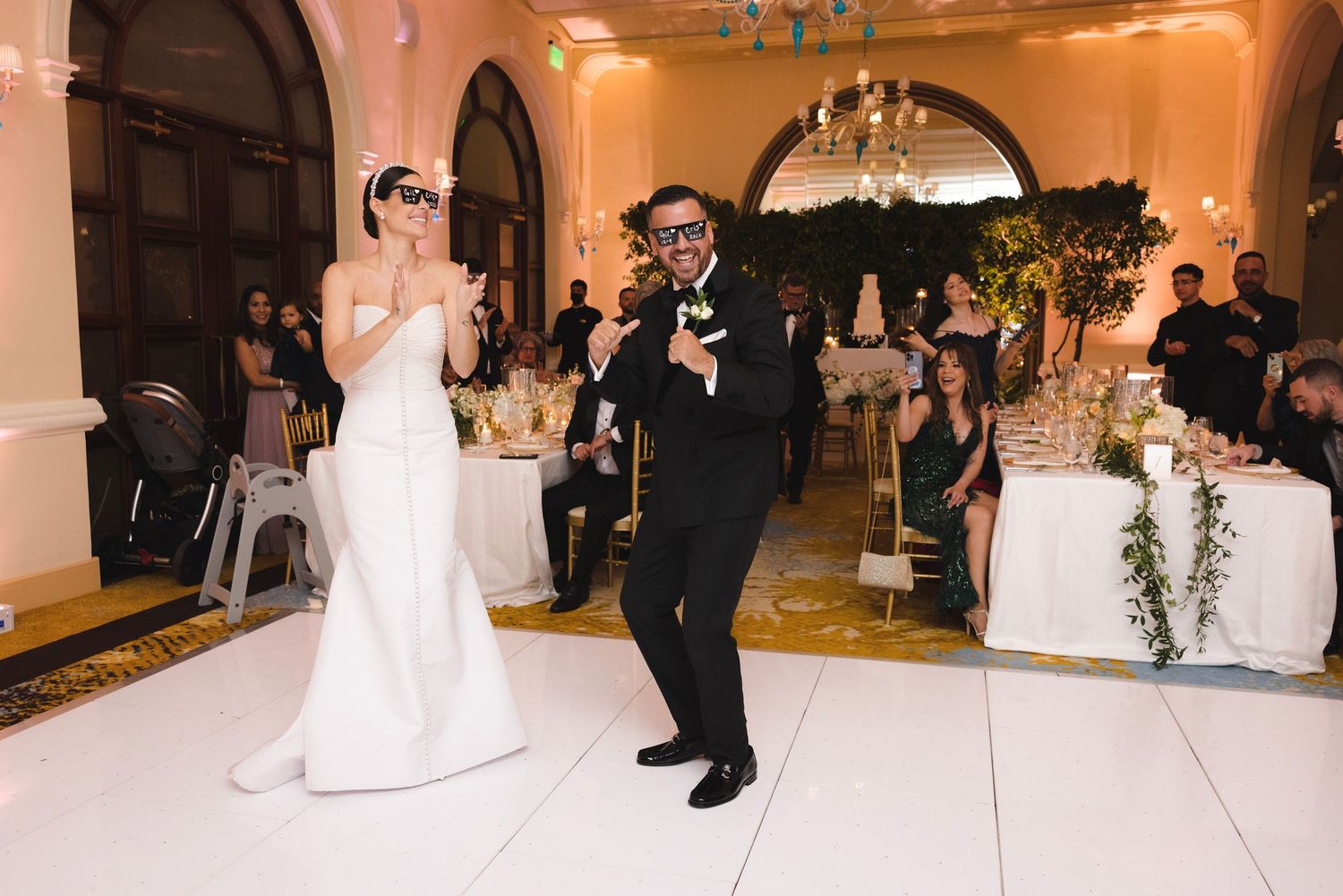 Two figures dancing together on a white dance floor during a formal wedding reception in an elegant ballroom.