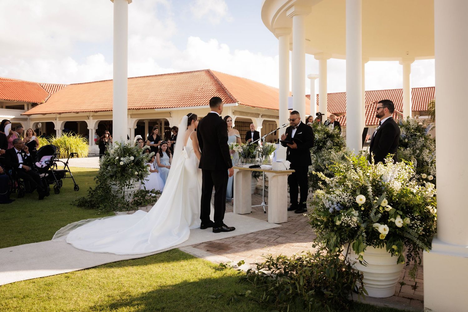 A romantic wedding ceremony setup under a white columned pavilion with lush floral arrangements and greenery.