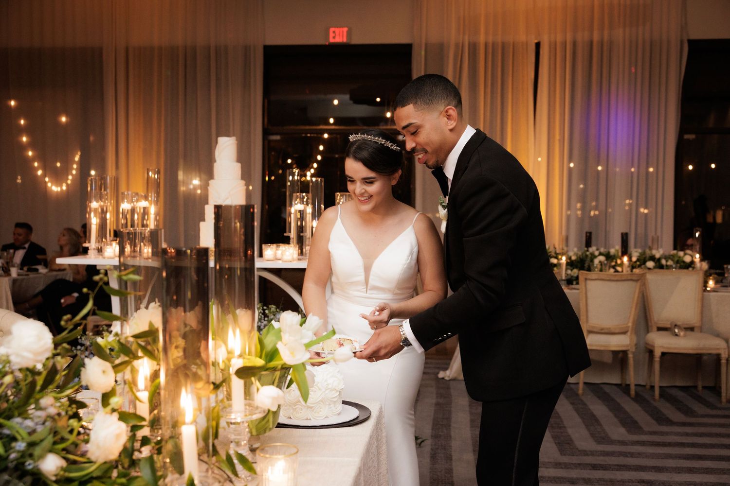 A couple cuts their elegant white wedding cake at an upscale reception venue with romantic lighting and floral decor.